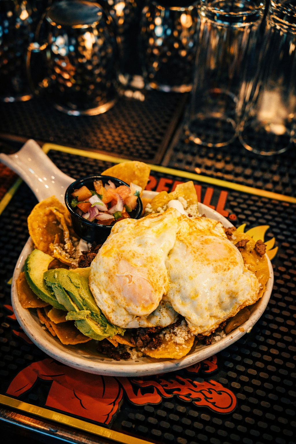 Bowl of Mexican food with nachos, fried eggs, avocado slices, and side of pico de gallo. In the background, glasses and bottles on a bar counter.
