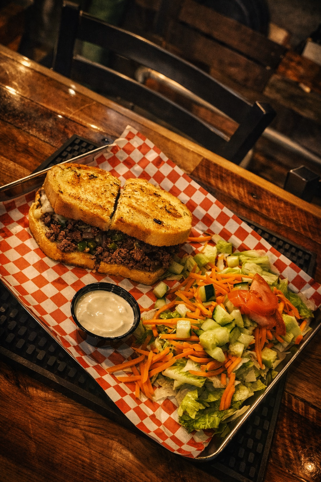 A sandwich with ground beef, lettuce, and cheese on toasted bread served with a side salad of lettuce, cucumbers, carrots, and a slice of tomato, along with a small cup of ranch dressing, all on a checkered paper-lined tray on a wooden table.