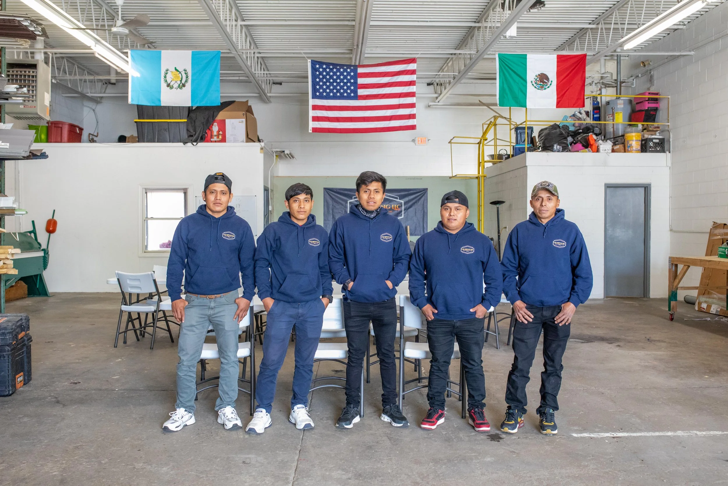 Five young men standing side by side dressed in blue hoodies inside a spacious warehouse with flags of Guatemala, the United States, and Mexico hanging from the ceiling.