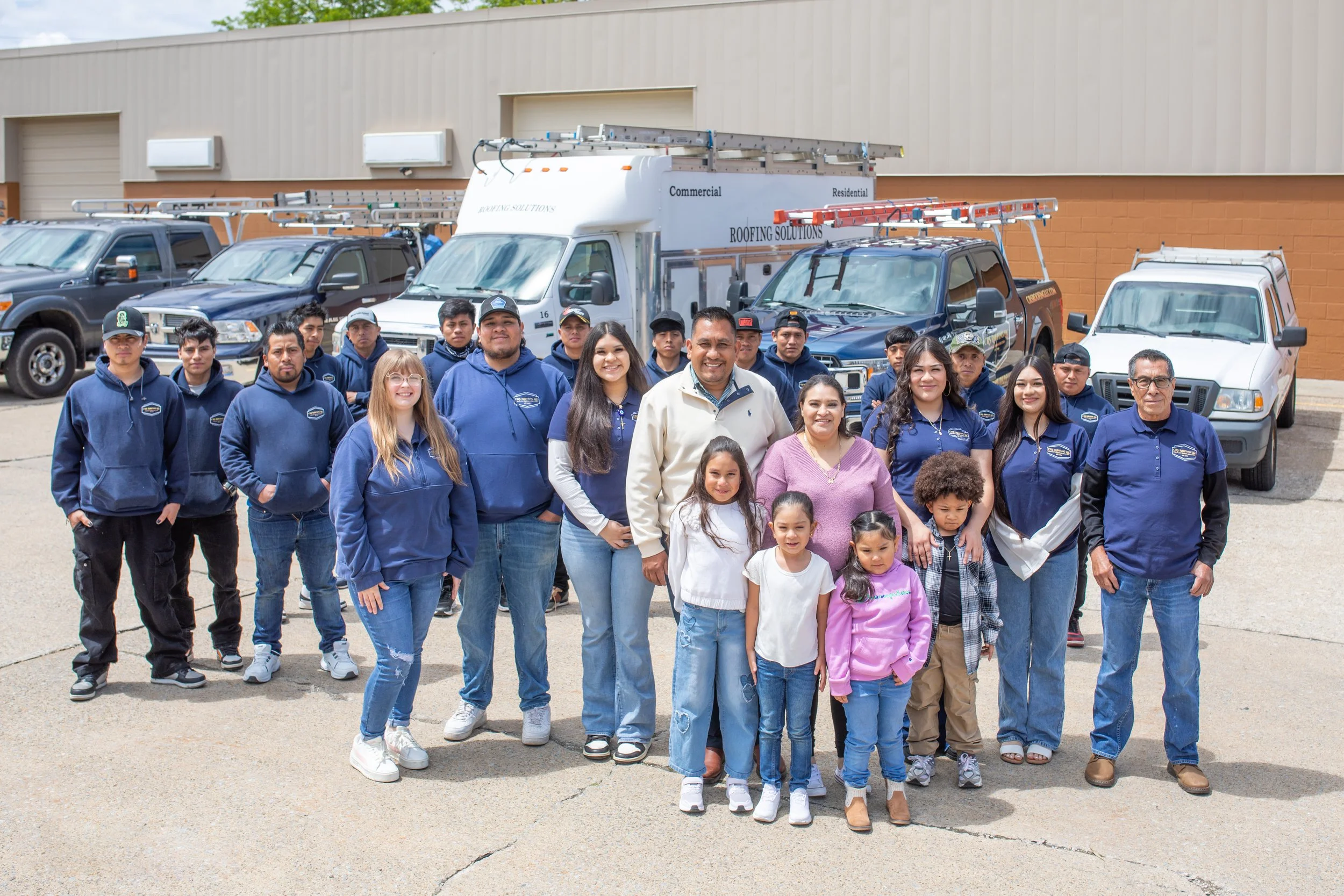 Group photo of adults and children standing in front of roofing and utility vehicles outside