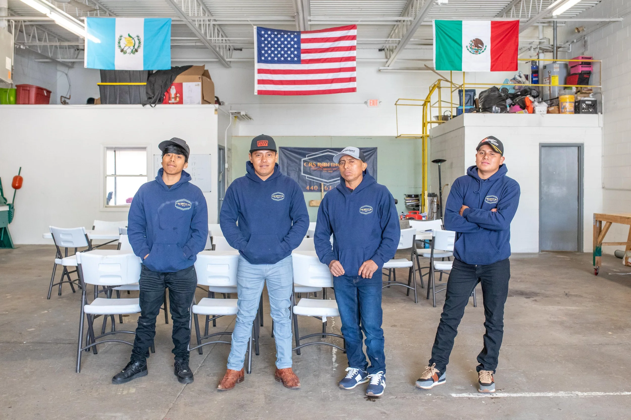 Four men standing in a workshop or garage, wearing matching blue hoodies with a logo, with flags of Guatemala, the United States, and Mexico hanging above them.