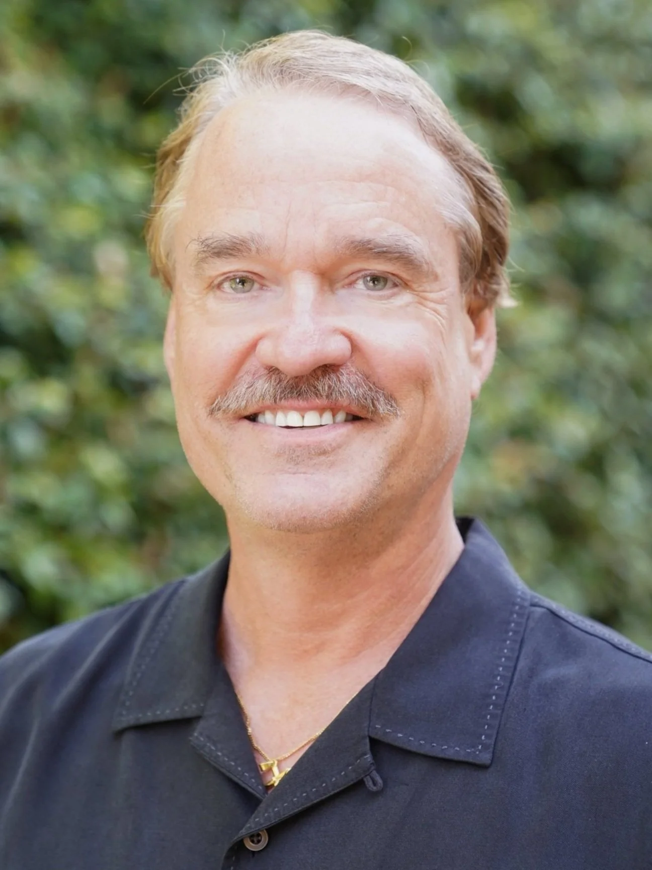 A smiling man with light brown hair and a mustache, wearing a dark shirt and a gold chain, standing outdoors with green foliage in the background.