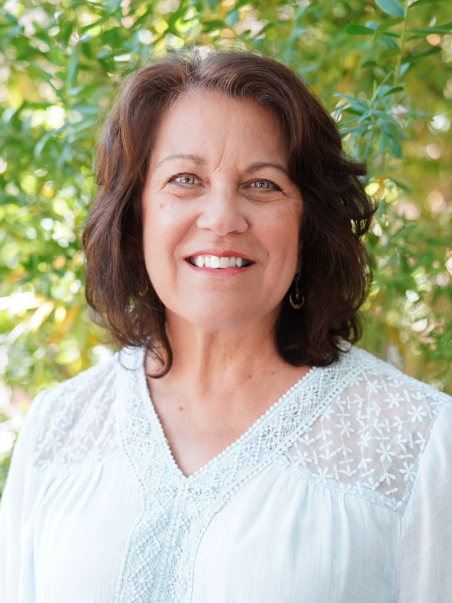 A middle-aged woman with shoulder-length dark hair and blue eyes, smiling while looking at the camera, outdoors with green leaves in the background, wearing a white embroidered blouse.