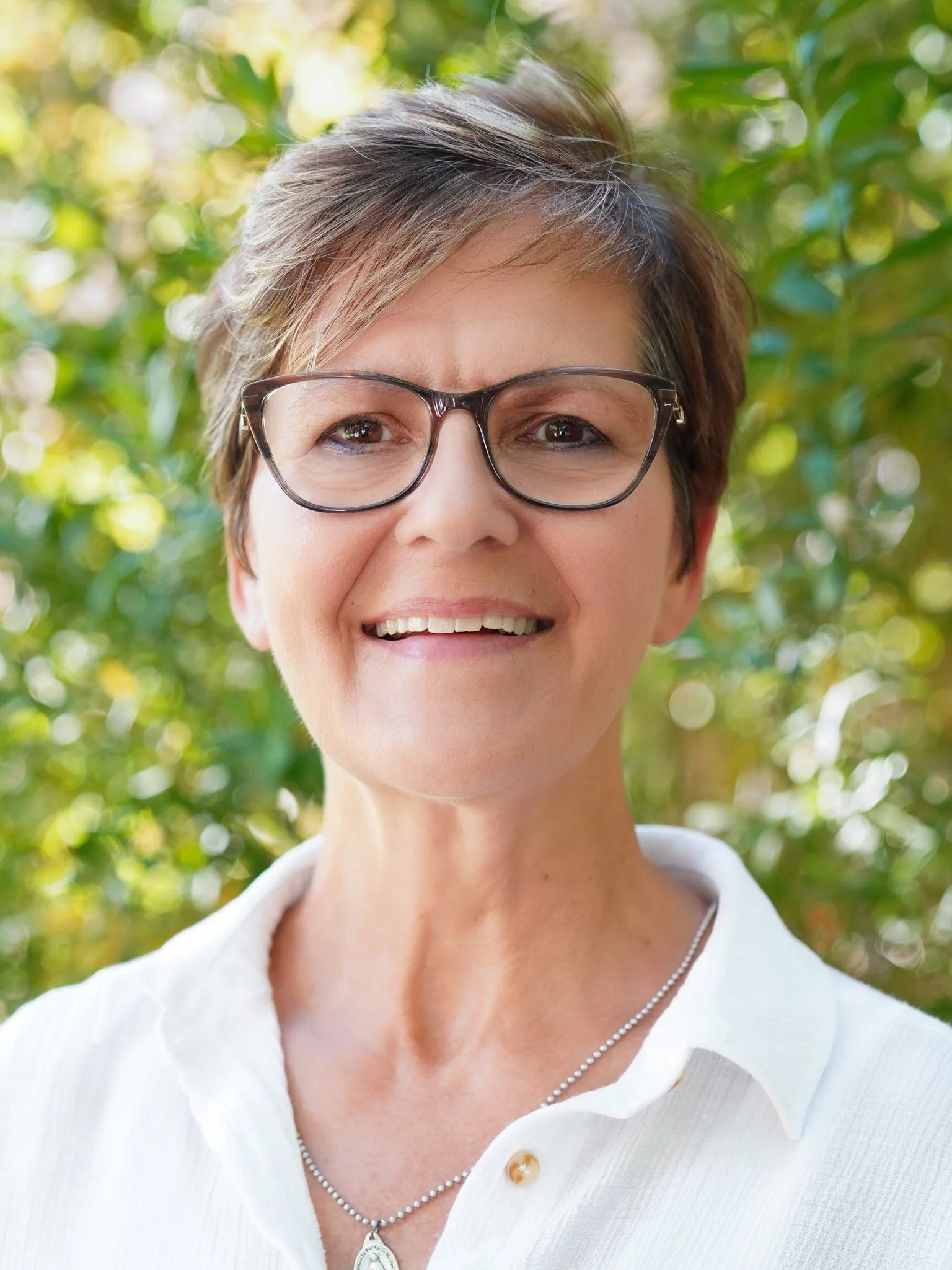Smiling middle-aged woman with short gray hair and glasses, wearing a white collared shirt, outdoors with green foliage background.