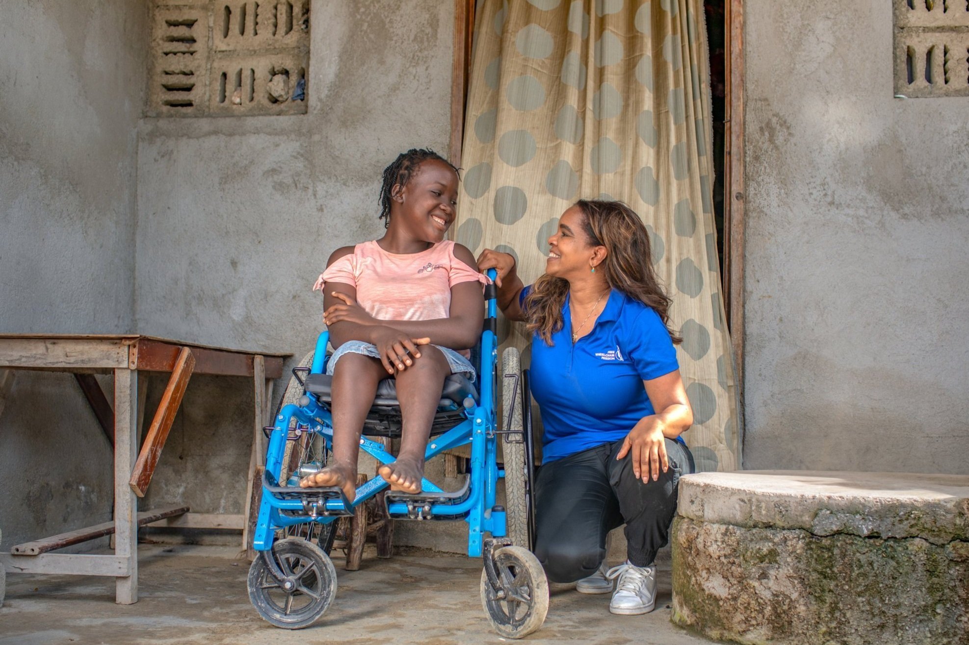 A young girl in a pink shirt sitting in a blue wheelchair, smiling, while a woman in a blue shirt kneels beside her, also smiling, outside a concrete building with a curtain in the background.