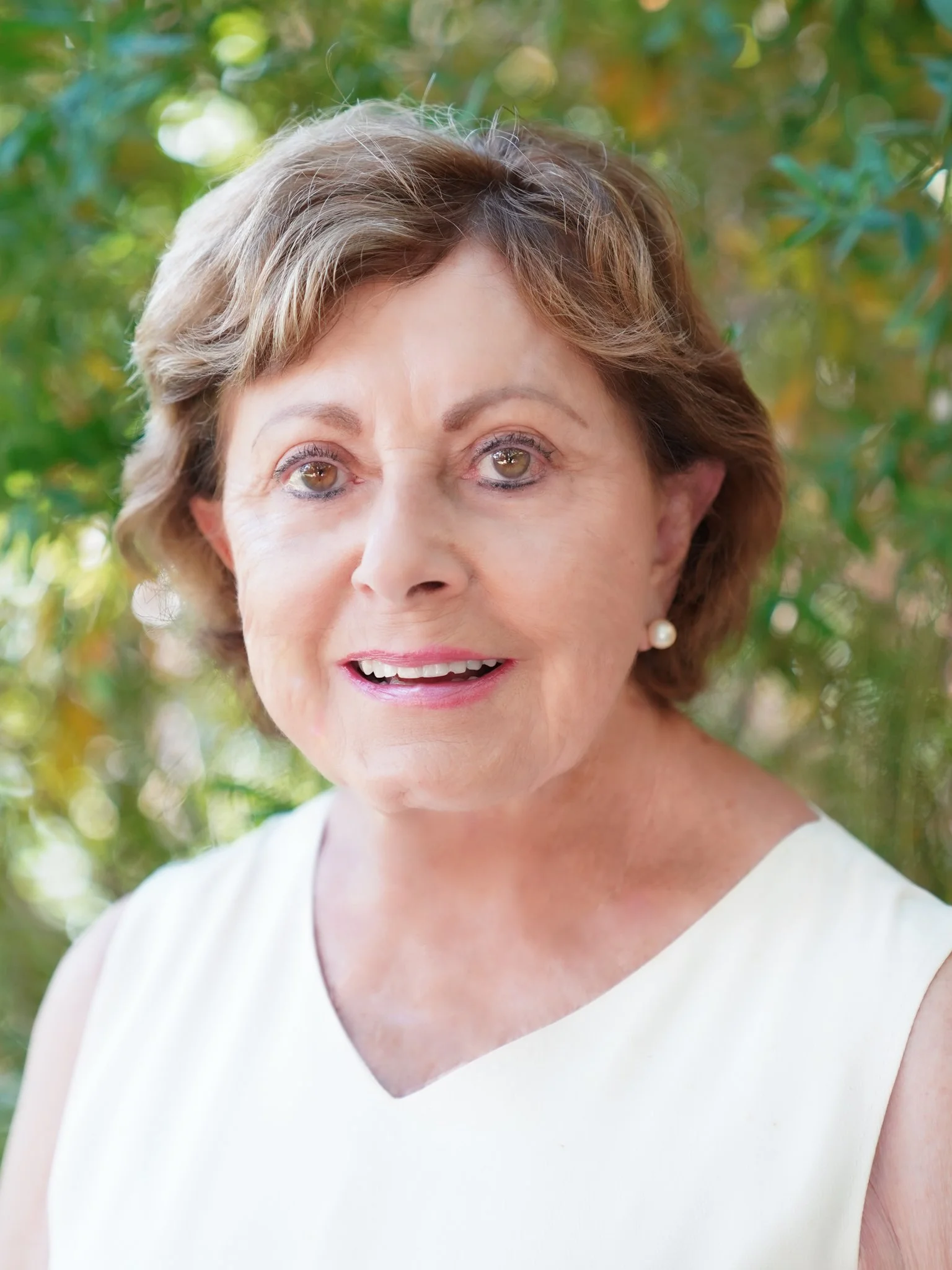 A close-up portrait of an elderly woman with short brown hair, wearing pearl earrings and a white sleeveless top, smiling softly, outdoors with green foliage in the background.