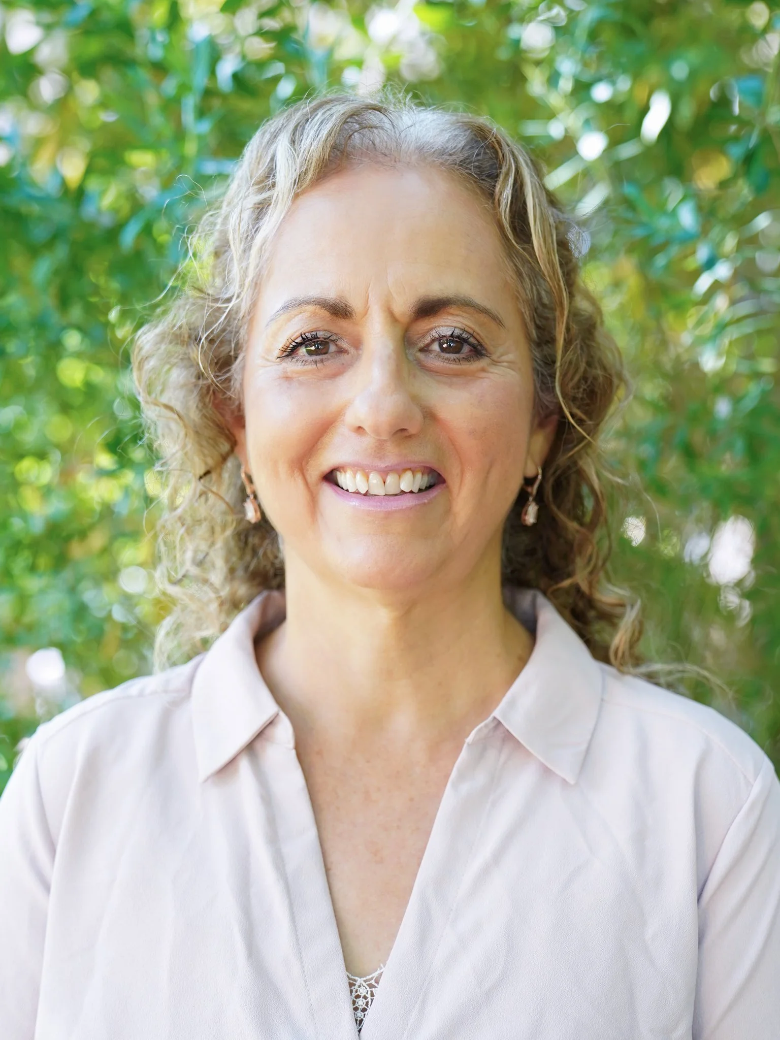 A woman with curly blonde hair smiling outdoors with green foliage in the background.