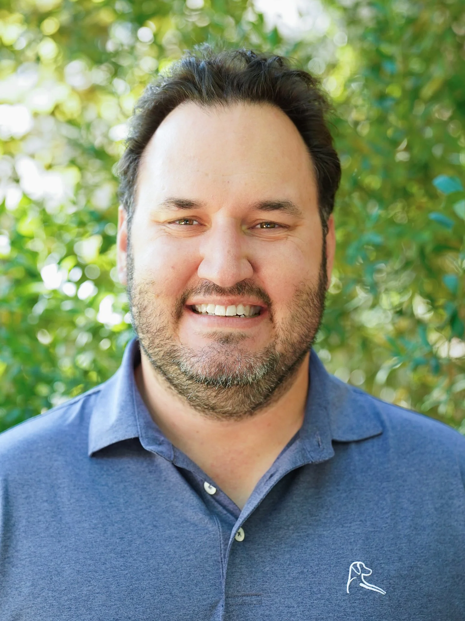 A man with dark, curly hair and a beard smiling, outdoors with green foliage in the background, wearing a blue polo shirt.