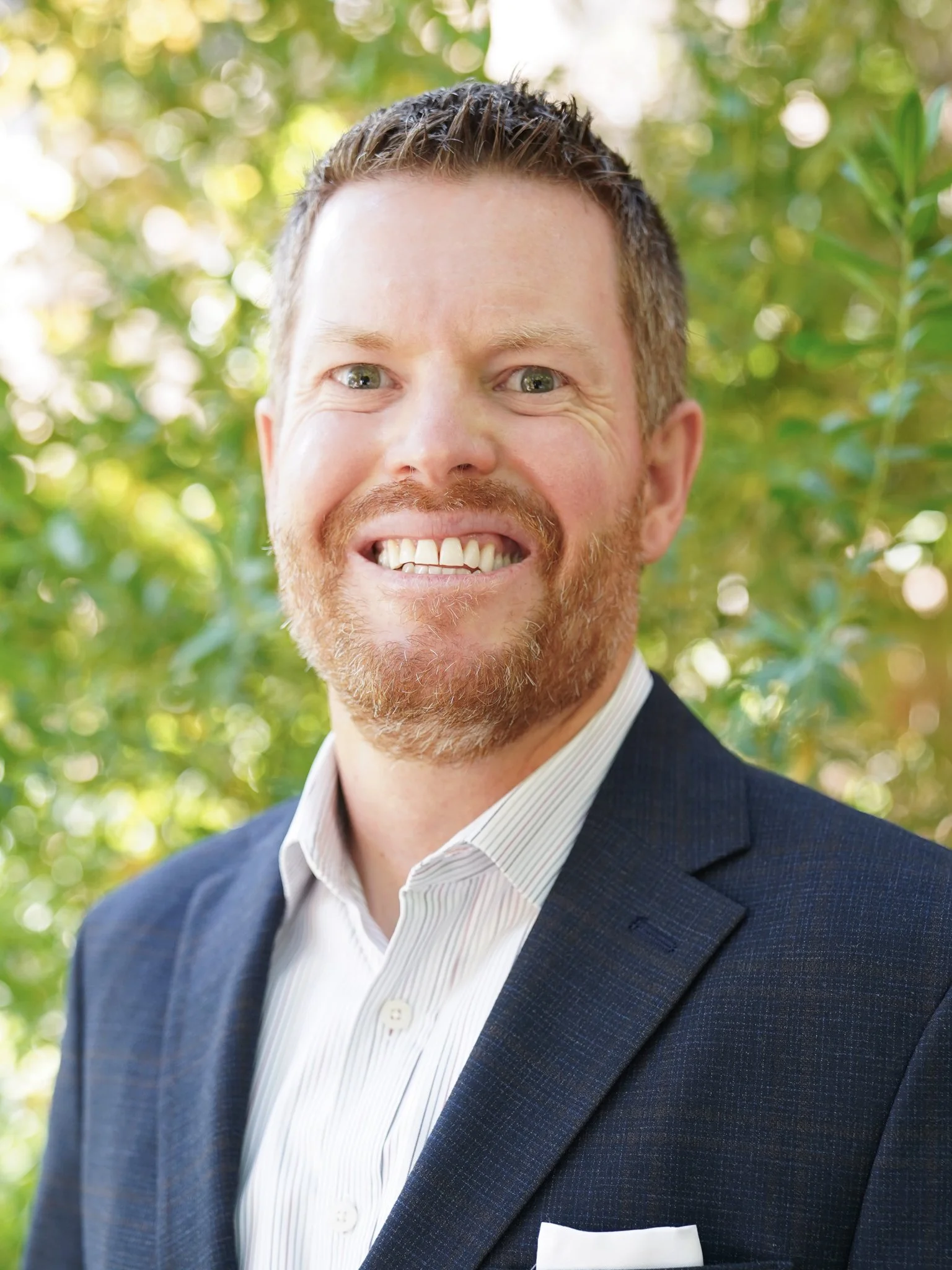 Business portrait of a smiling man with short brown hair and a beard, wearing a dark suit and white shirt, outdoors with green foliage in the background.