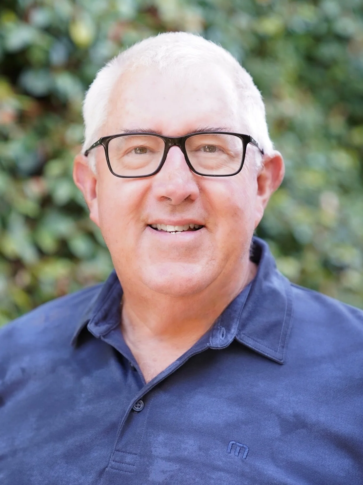 An older man with white hair and glasses, wearing a dark blue shirt, smiling outdoors with green foliage in the background.