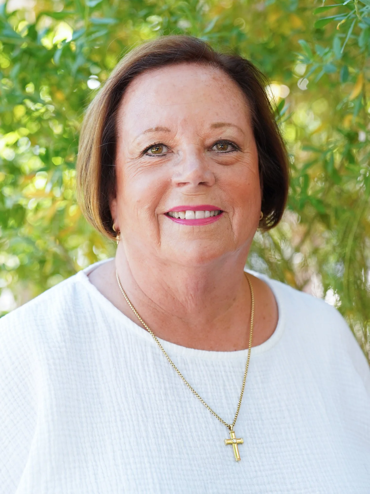 A middle-aged woman with short brown hair, wearing a white top and a gold cross necklace, smiling outdoors with green trees in the background.