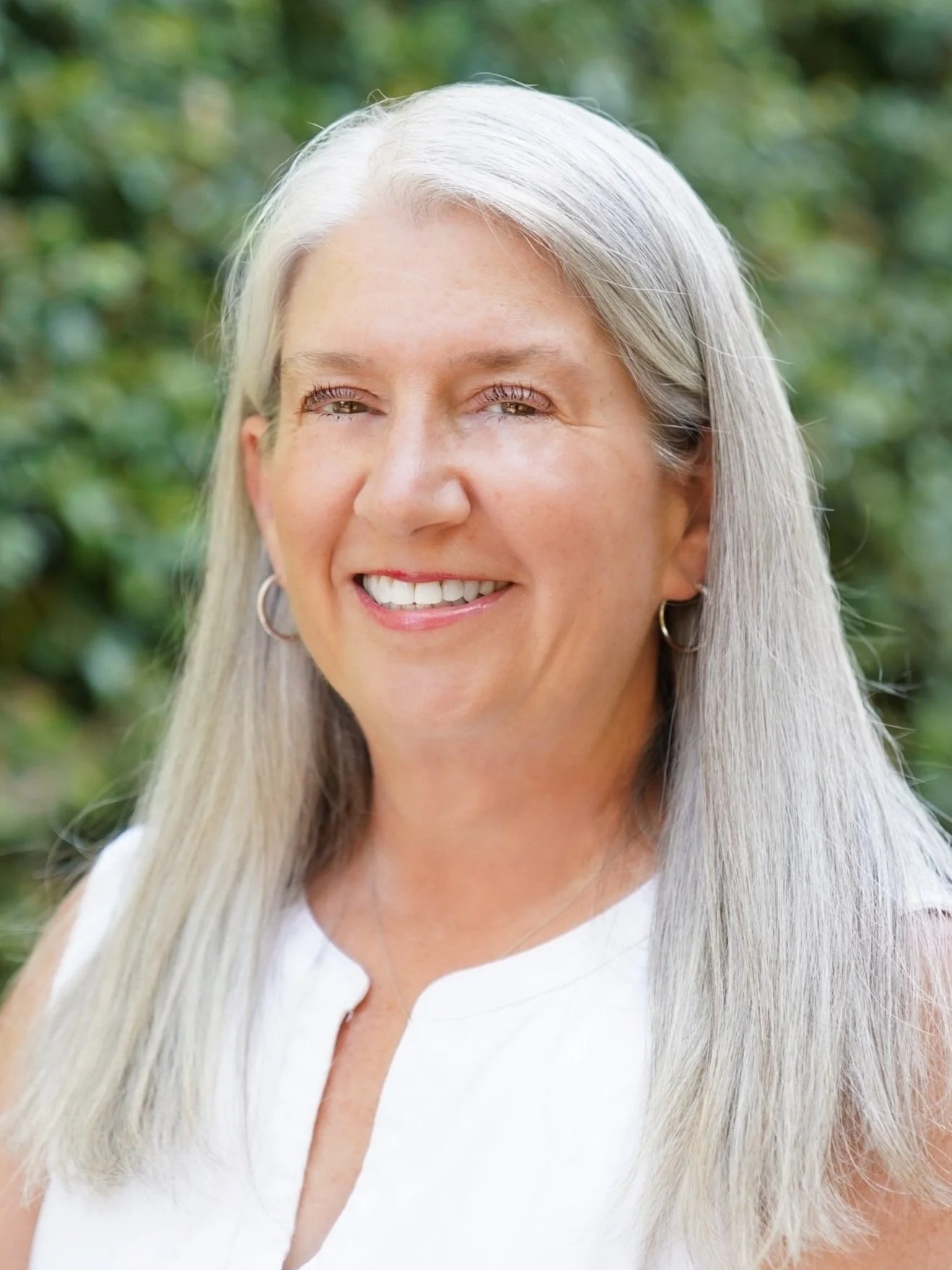 A smiling woman with long silver hair and light skin, wearing a white sleeveless top and hoop earrings, outdoors with a blurred green natural background.