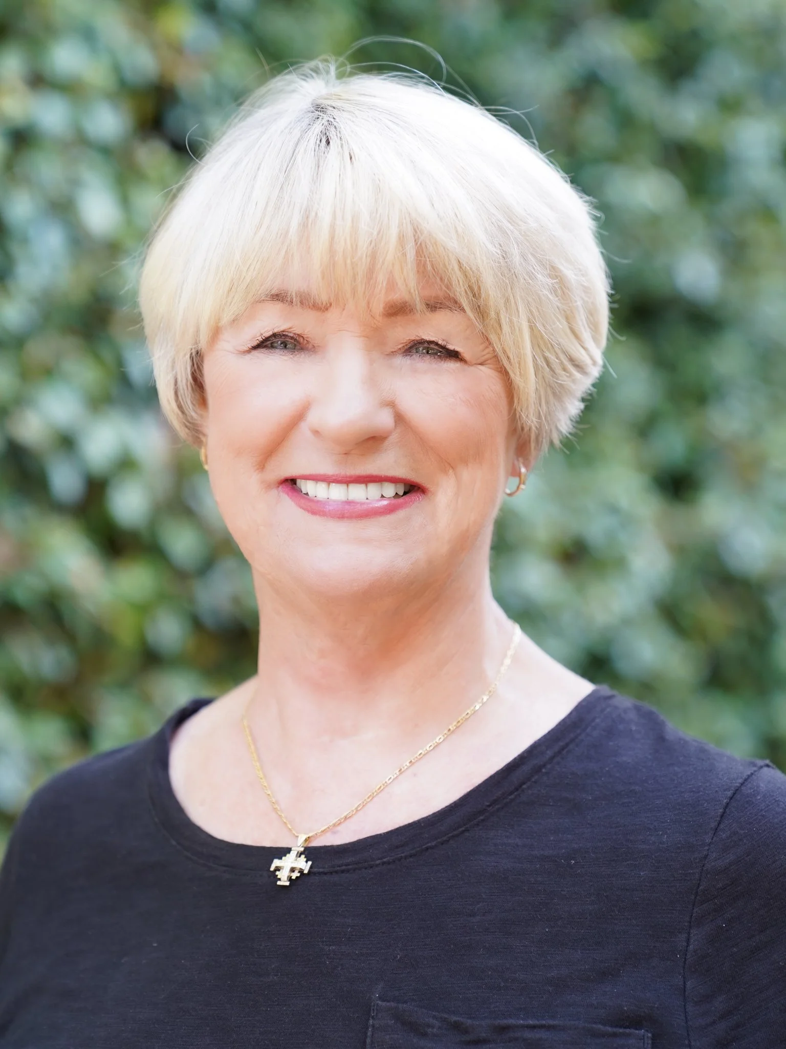 A smiling middle-aged woman with short blonde hair wearing a black top and a gold necklace with a cross pendant, standing outdoors with a blurred green leafy background.