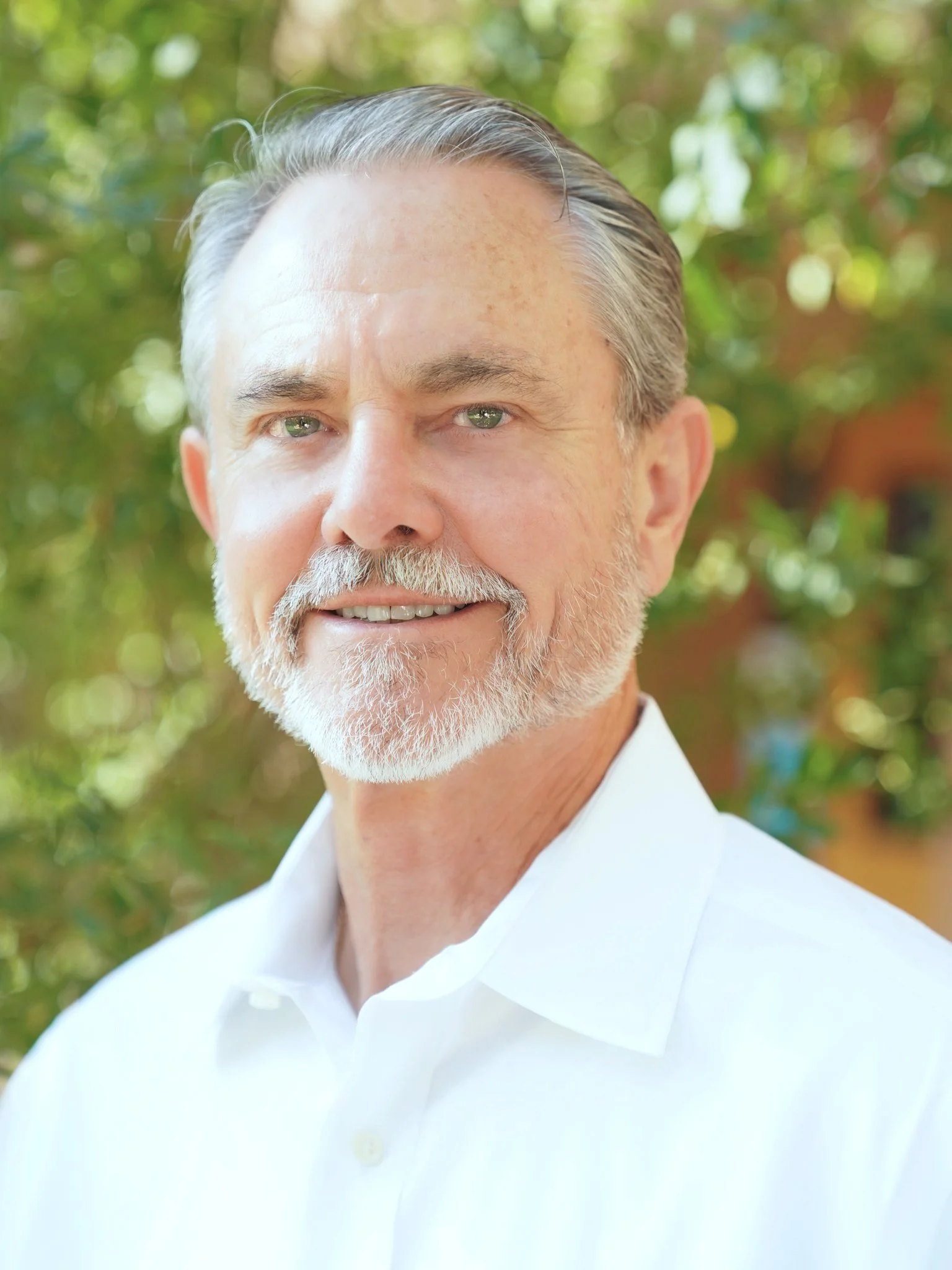 Close-up portrait of a smiling older man with gray hair and beard, wearing a white dress shirt, outdoors with green foliage in the background.