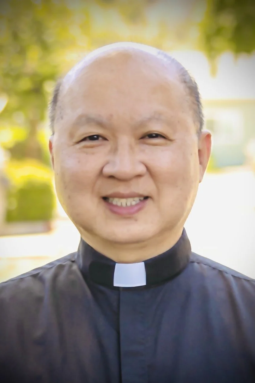 Portrait of a middle-aged man wearing glasses and a black clerical shirt with a white clerical collar, standing outdoors with green foliage in the background.