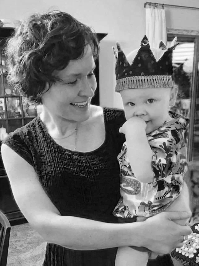 A woman holding a young boy wearing a birthday crown, inside a home with a window and curtains in the background.