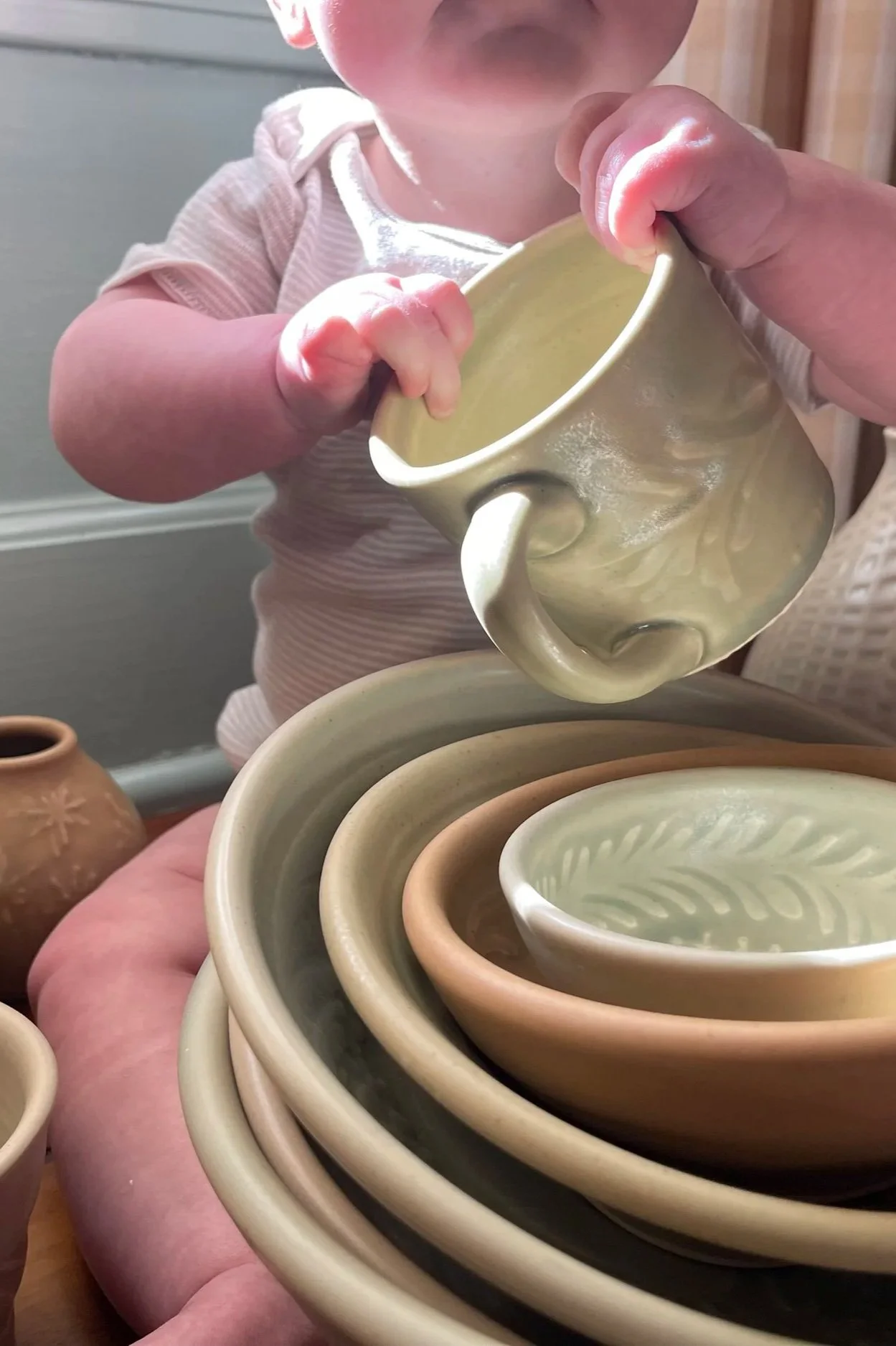 A child's hand holding a ceramic mug over a stack of similar mugs with another person nearby, with additional pottery in the background.