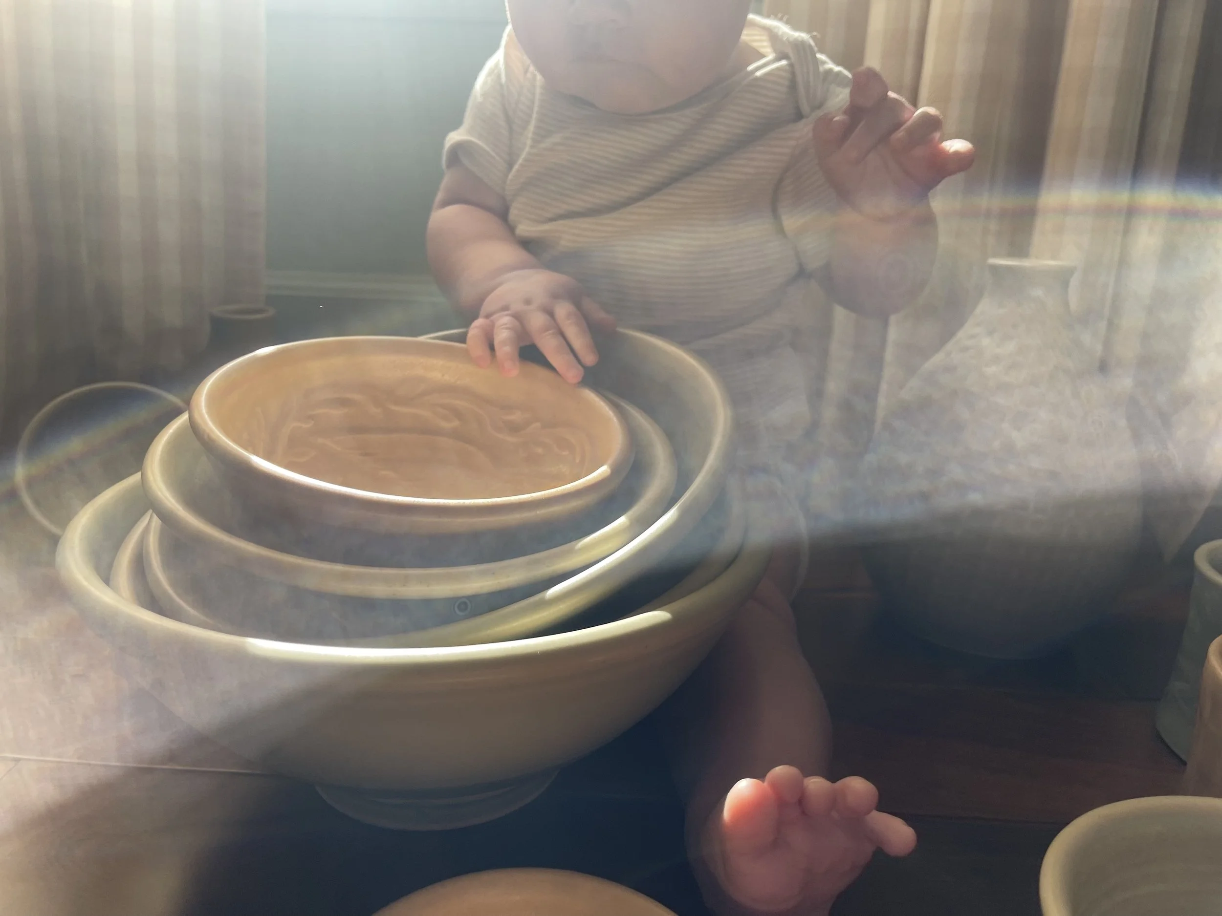 A child with a striped shirt is reaching toward the camera, with a bowl of soup and multiple stacked bowls nearby, inside a room with striped curtains and sunlight streaming in.