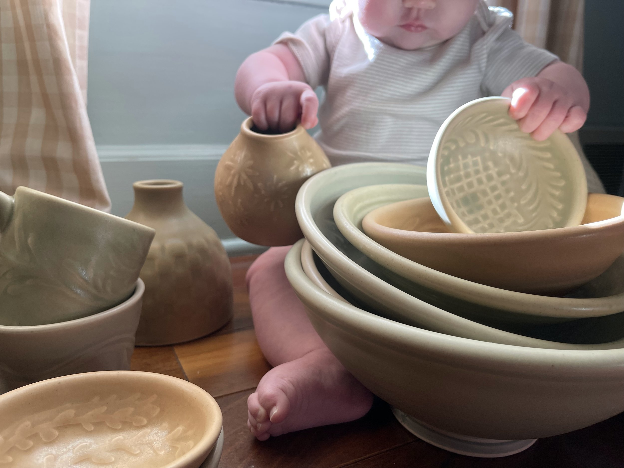 A young child with chubby cheeks and wearing a beige striped onsie stacking mineral colored ceramic bowls on a wooden floor surrounded by various pottery pieces.