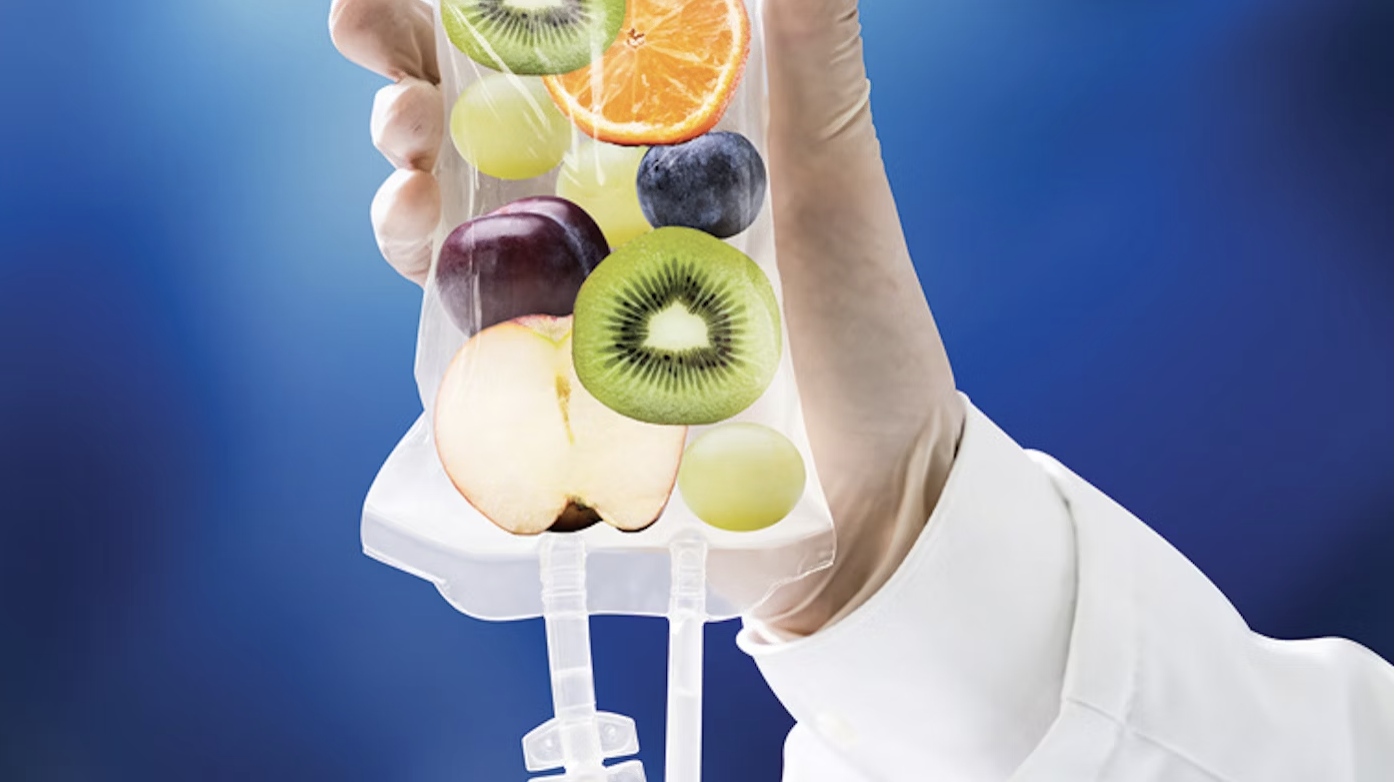 A hand holding an IV bag filled with various fruit slices, including orange, kiwi, apple, grapes, and a plum, against a blue background.