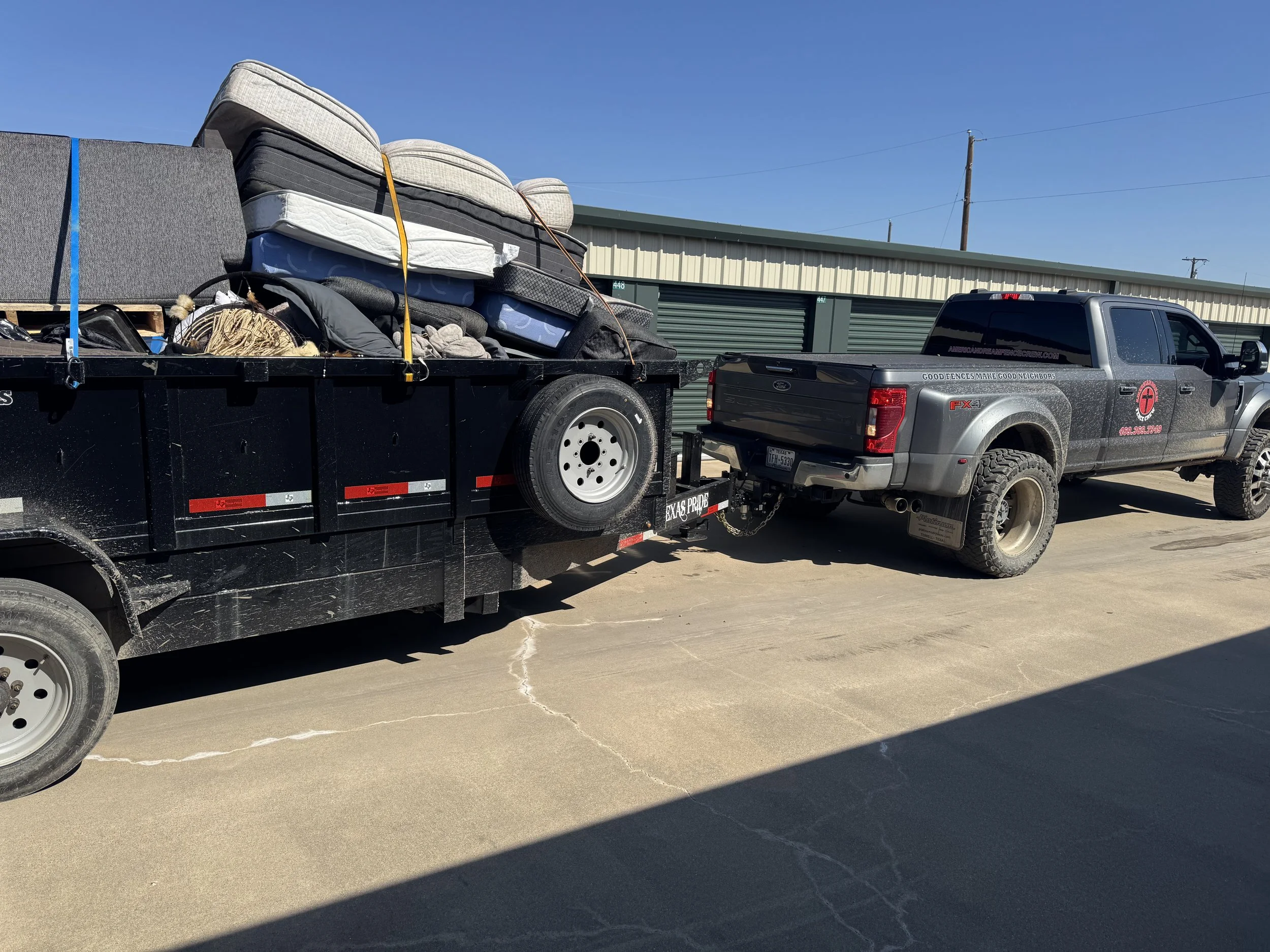 A black flatbed trailer attached to a gray Ford F-150 pickup truck parked in front of storage units under a clear blue sky. The trailer is loaded with several mattresses and other household items secured with straps.