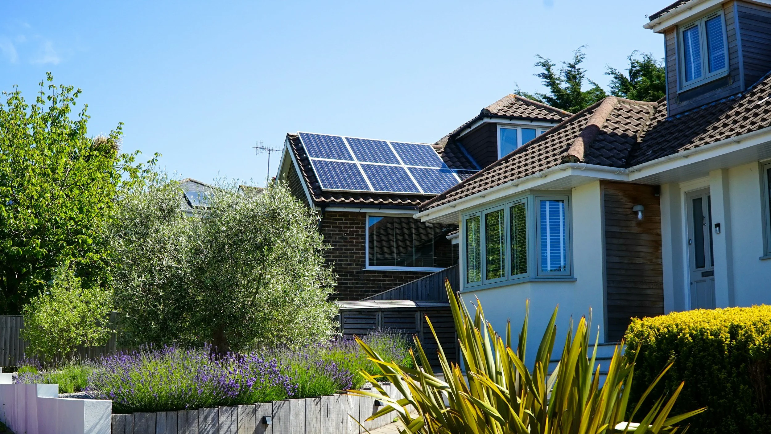 Residential house with solar panels on the roof, surrounded by landscaped garden with green trees, purple flowers, and yellow bushes, under a clear blue sky.