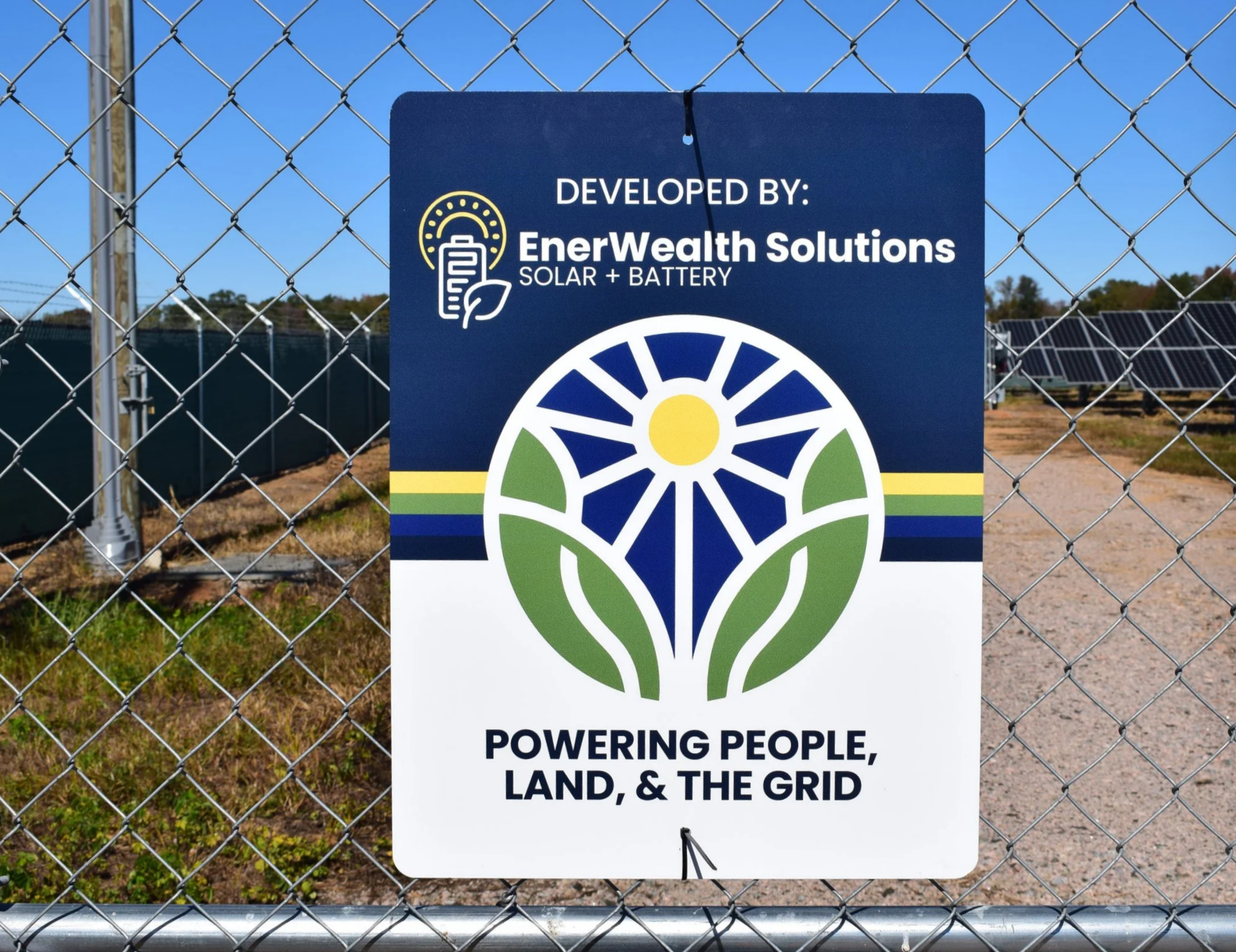 Sign on chain-link fence for EnerWealth Solutions, with a logo featuring a stylized flower and leaves, and solar panels in the background. The sign states 'Developed by: EnerWealth Solutions Solar + Battery' and 'Powering people, land, & the grid.'