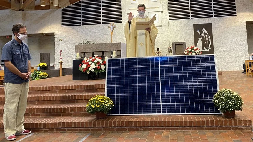A person in a blue shirt and khakis is standing with hands together in front of a priest at an altar inside a church, with a large solar panel and potted flowers in the foreground. The priest, dressed in white and wearing a face mask, is reading from