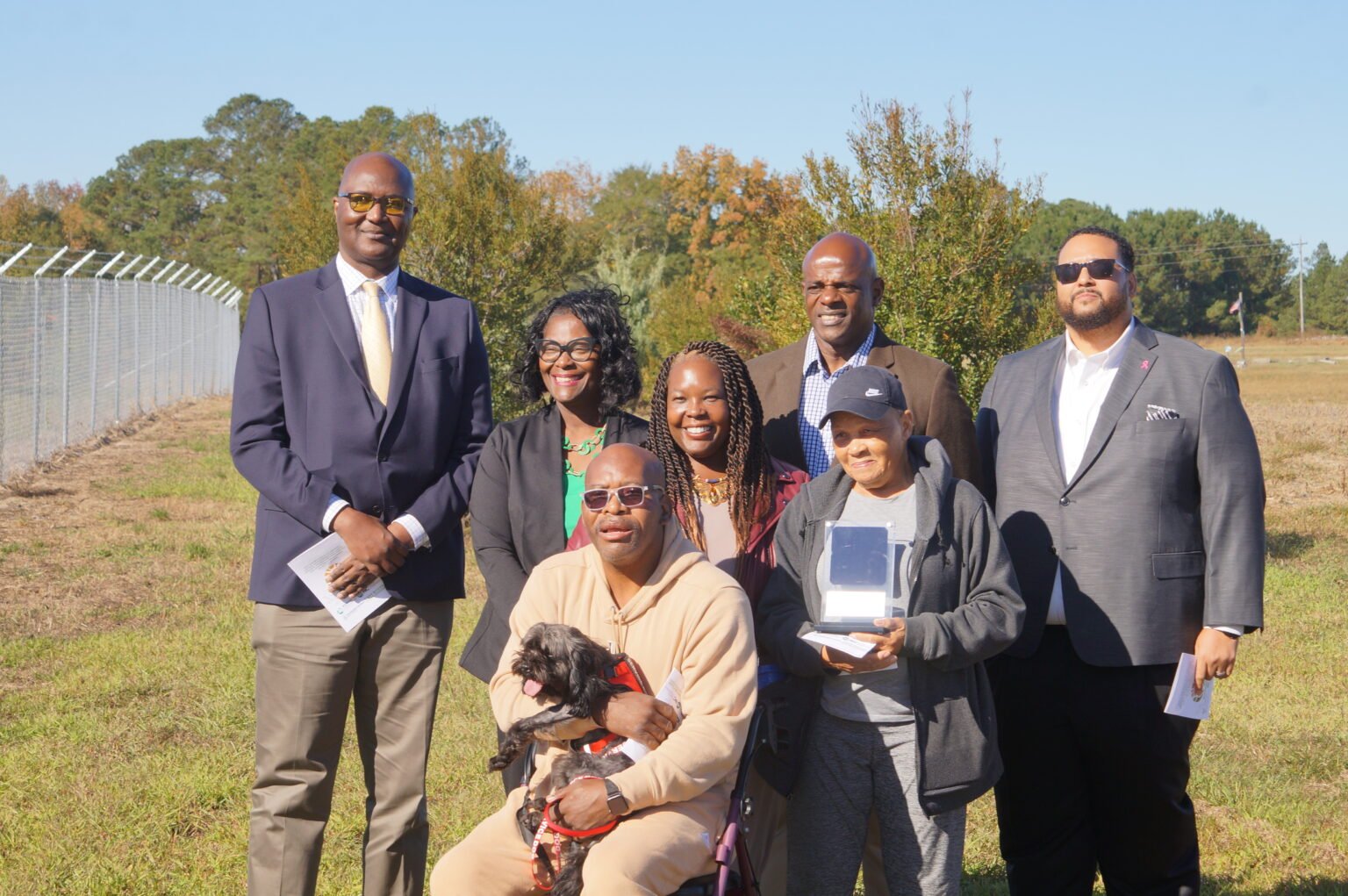 A group of adults, including a woman in a chair with a dog, standing outdoors on a sunny day with trees and a fence in the background.