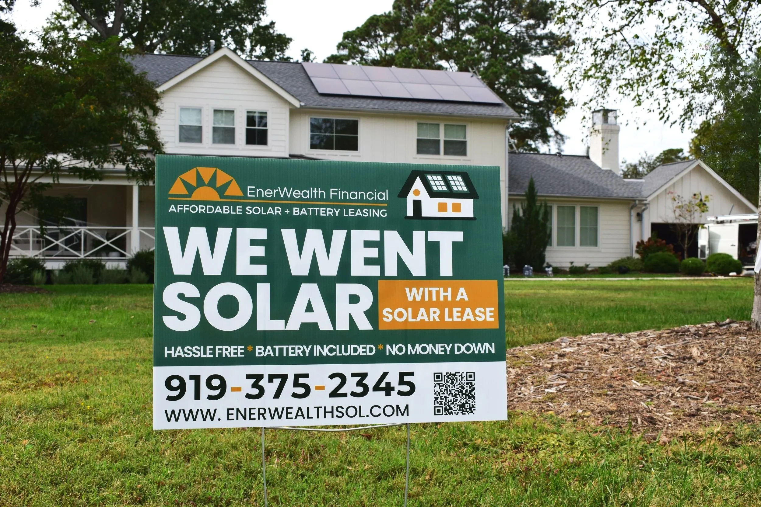 A yard sign in front of a white house with solar panels on the roof, advertising Solar leasing services by EnerWealth , mentioning hassle-free, battery included, no money down, with a phone number and website.