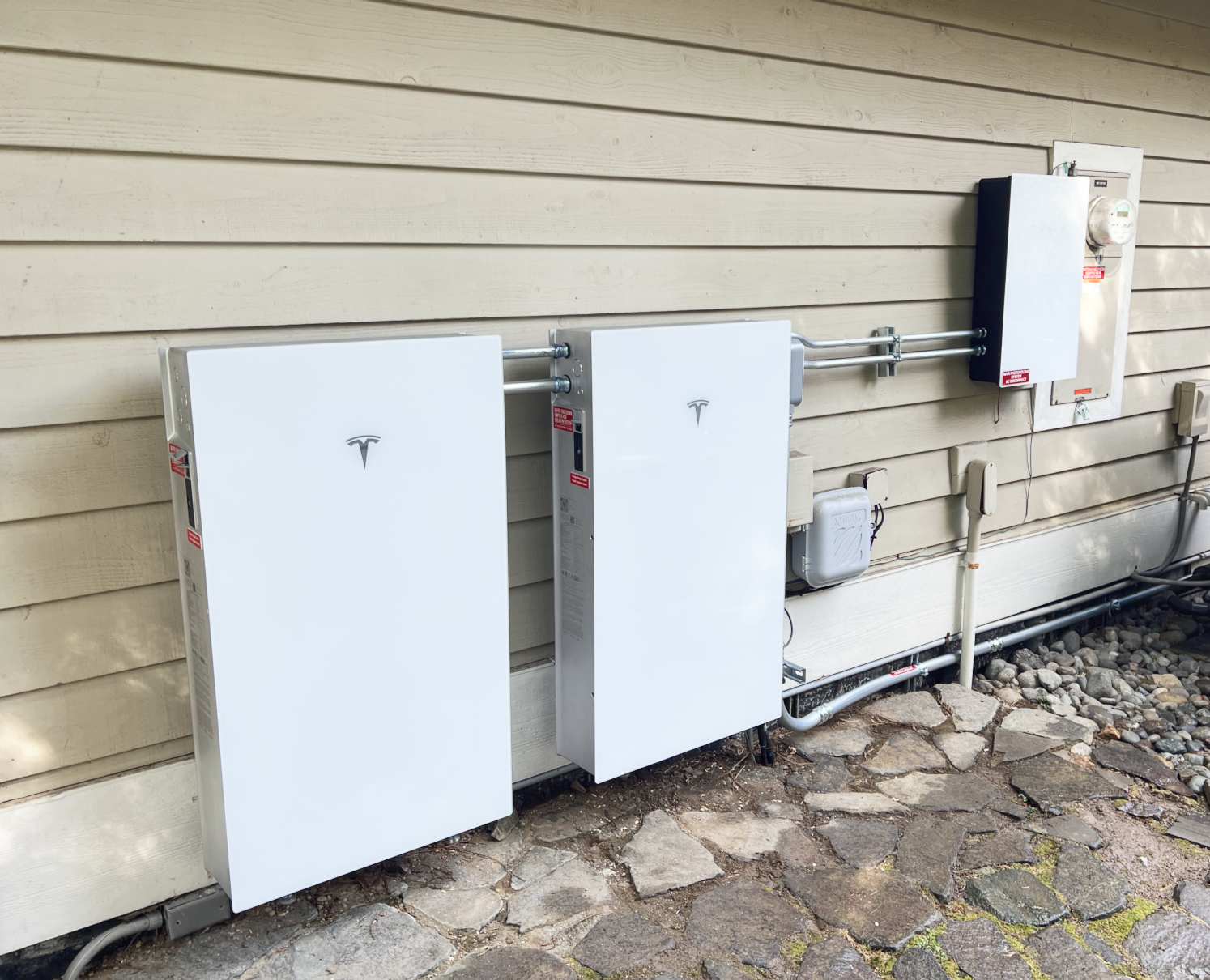 Outside view of a house with electrical equipment, including two white Tesla power walls, an electrical panel, and various wiring on a beige wooden siding wall, with a stone pathway in the foreground.