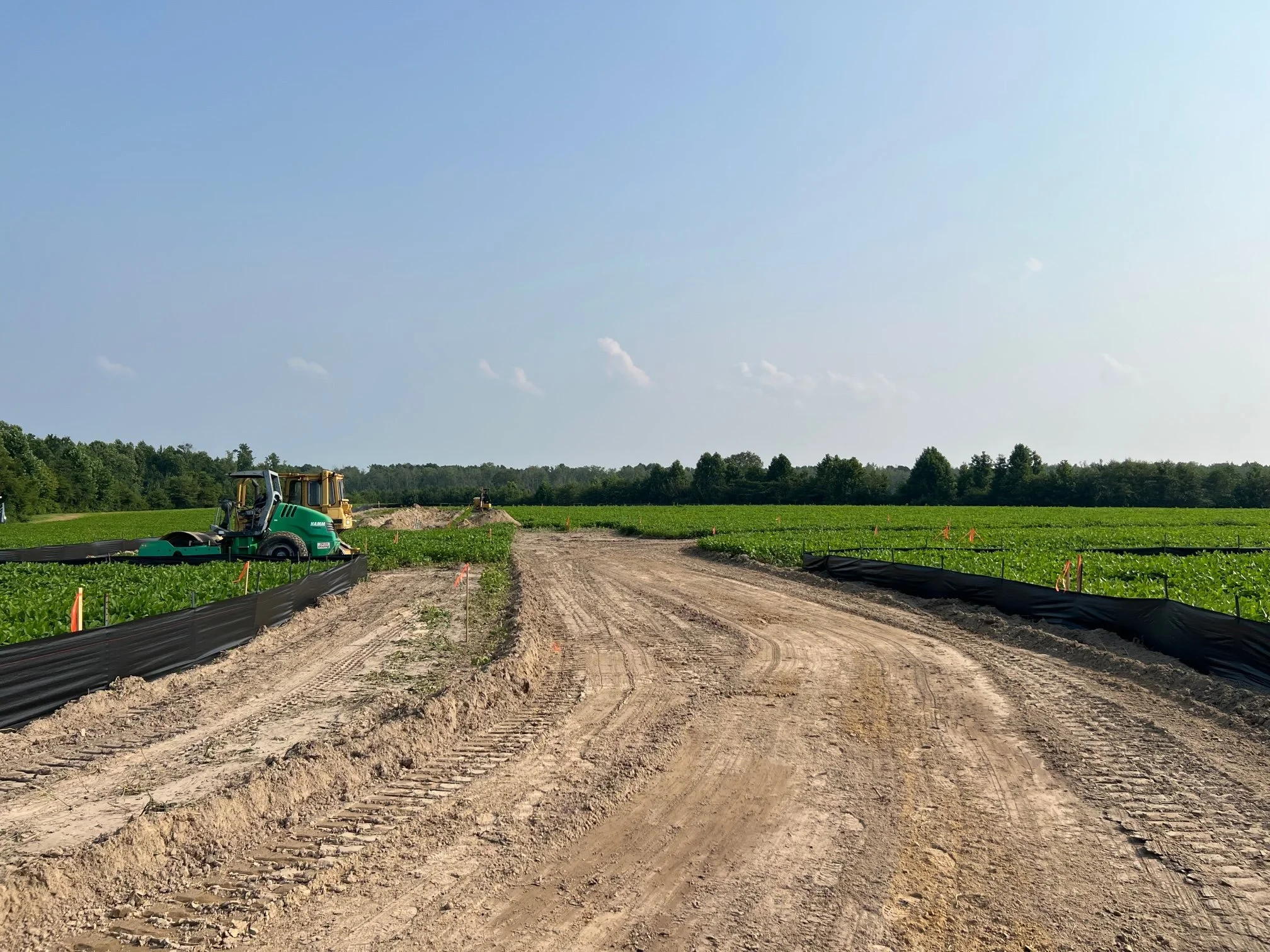 A rural construction site with a dirt road, agricultural fields on either side, and construction machinery under a clear blue sky.