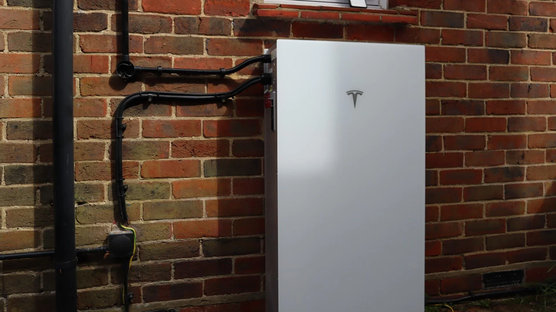 Electric vehicle charging station mounted on a brick wall with black conduits running into it, featuring the Tesla logo.