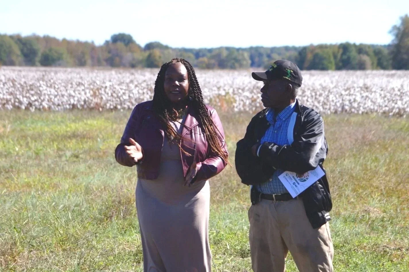 A woman and a man standing in a grassy field with a cotton field in the background during daytime.