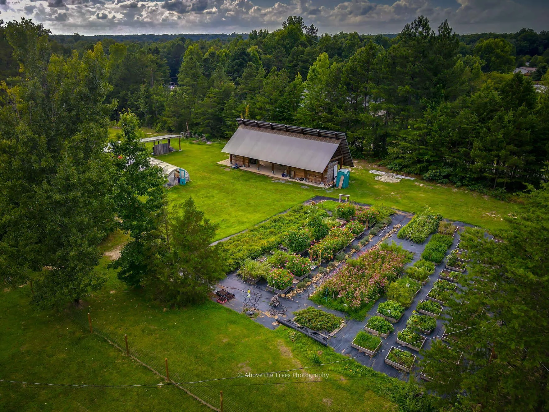 Aerial view of a lush, green farm with a large right-angle barn, vegetable garden beds, and a greenhouse, surrounded by trees under a cloudy sky.