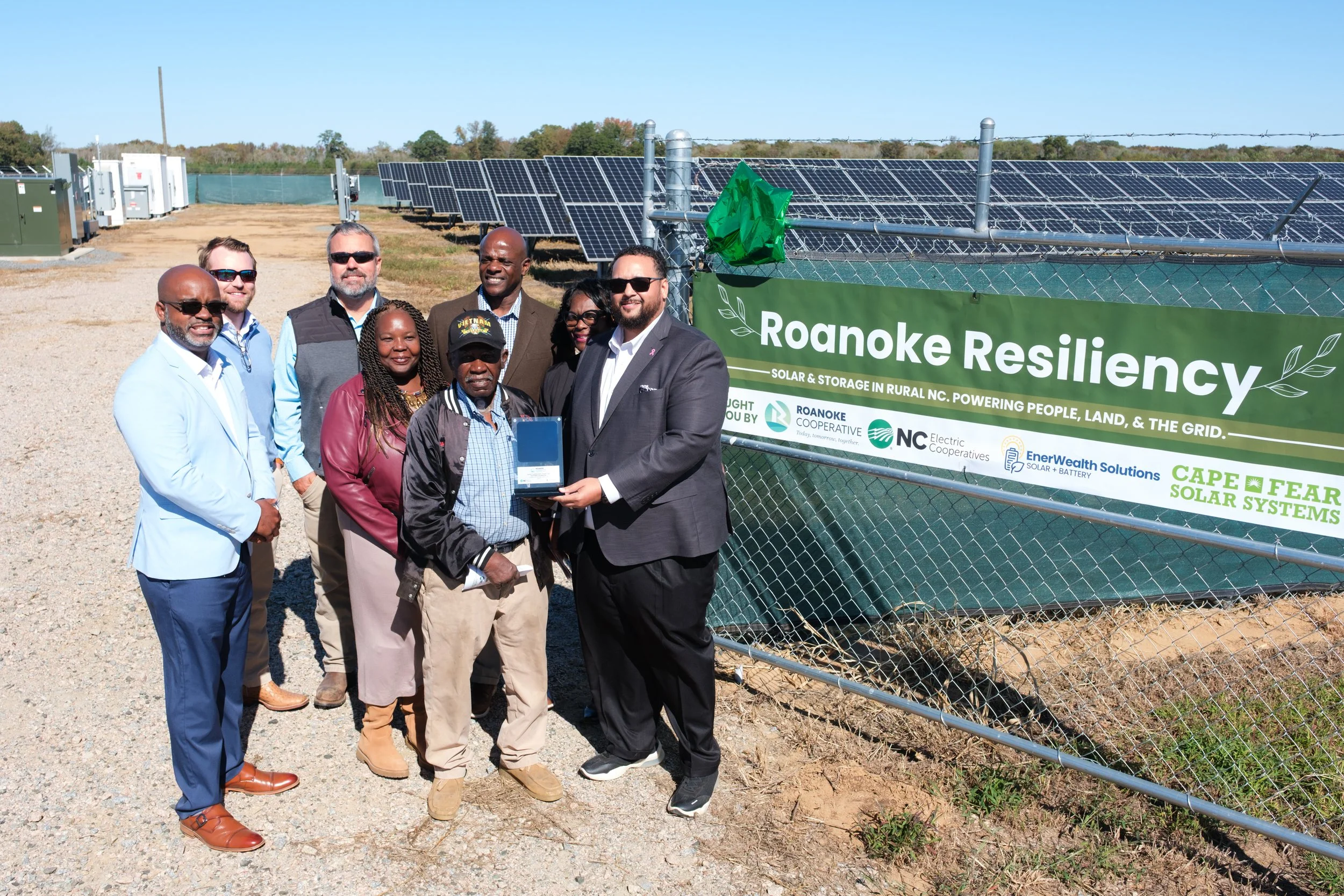 Group of people standing in front of solar panels and a sign that reads "Roanoke Resiliency: Solar & Storage in Rural NC. Powering People, Land, & The Grid."