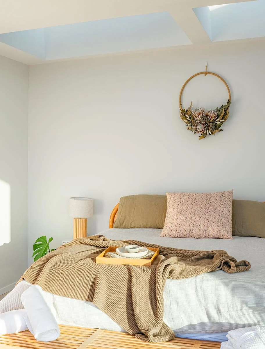 Bedroom with beige bed, floral pillow, tan and beige blankets, white tray with dishes, plant, lamp, and decorative flower hoop on white wall near skylight.