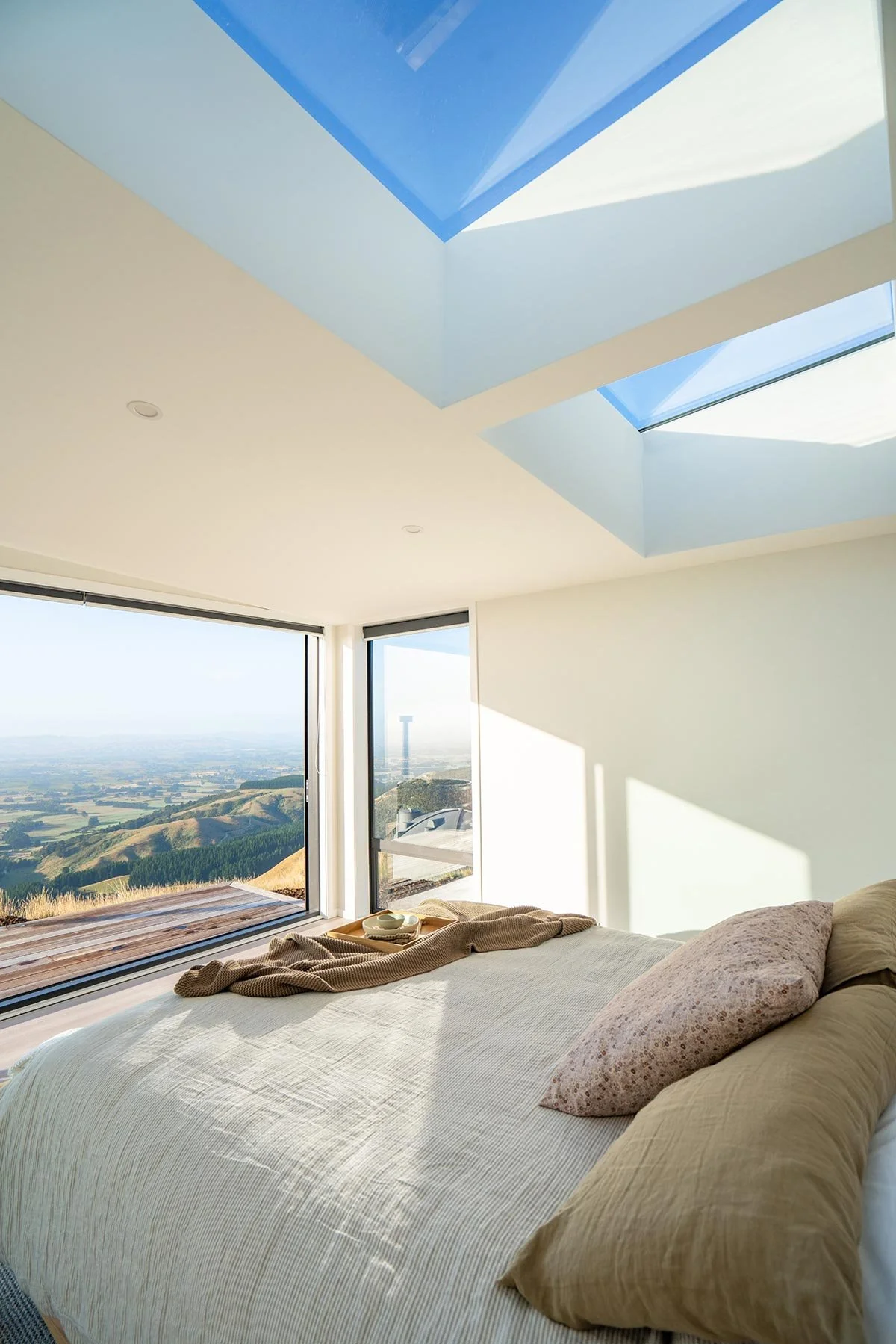 Bright bedroom with large windows showcasing a scenic landscape of rolling hills and fields, sunlit with skylights in the ceiling.
