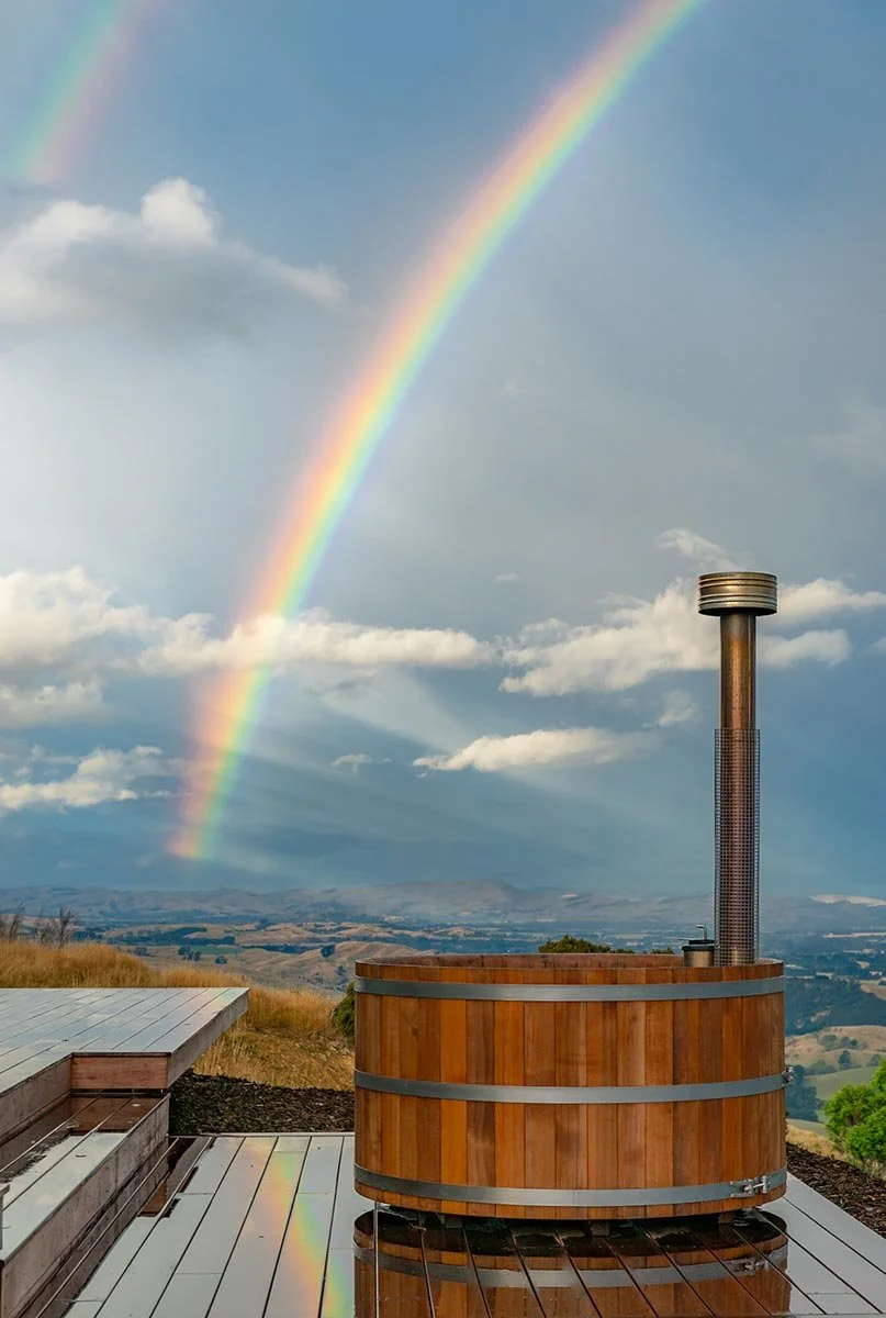 A rainbow over a wooden hot tub outdoors with a scenic landscape and cloudy sky in the background