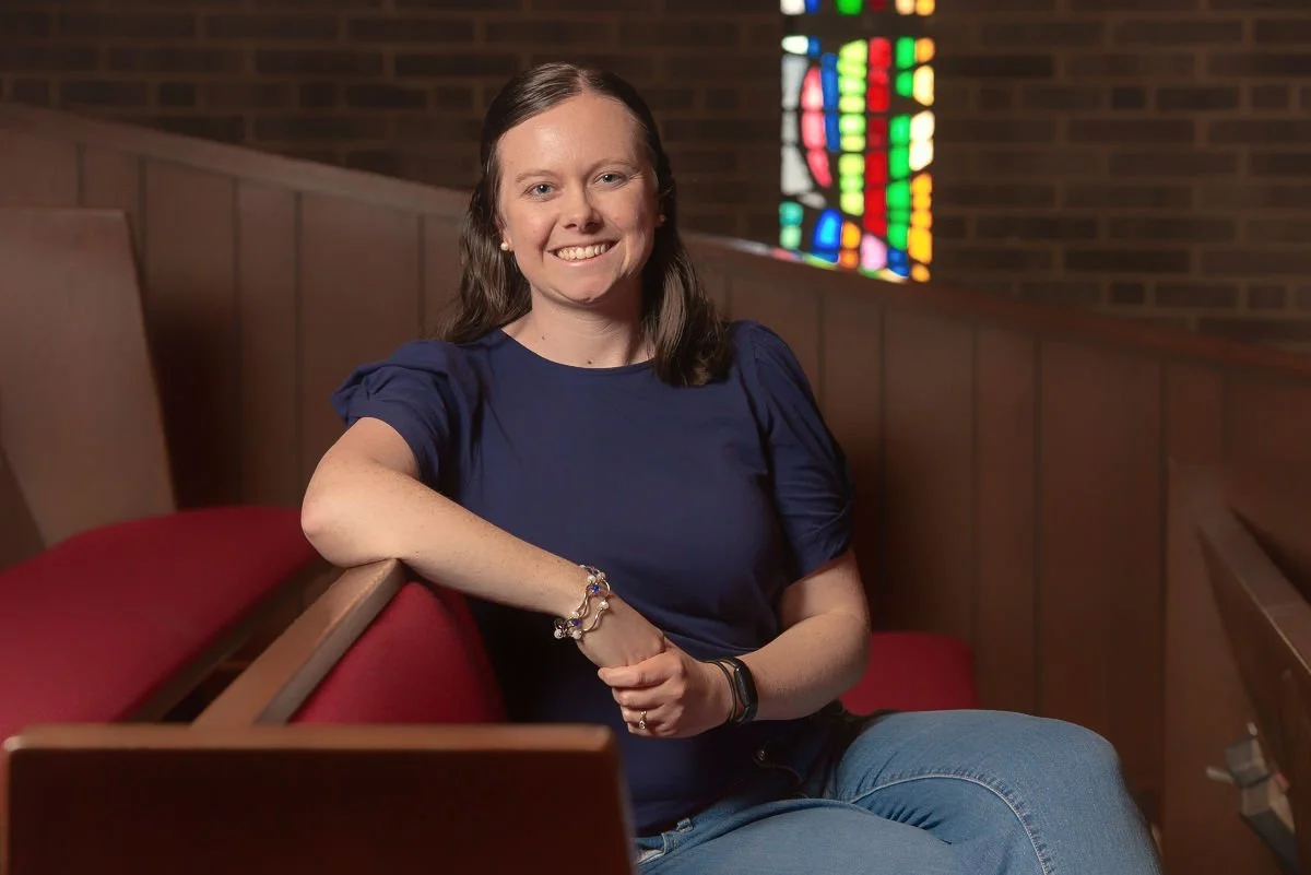 Person sitting in a church pew with a stained glass window in the background, wearing a navy blue shirt and jeans.
