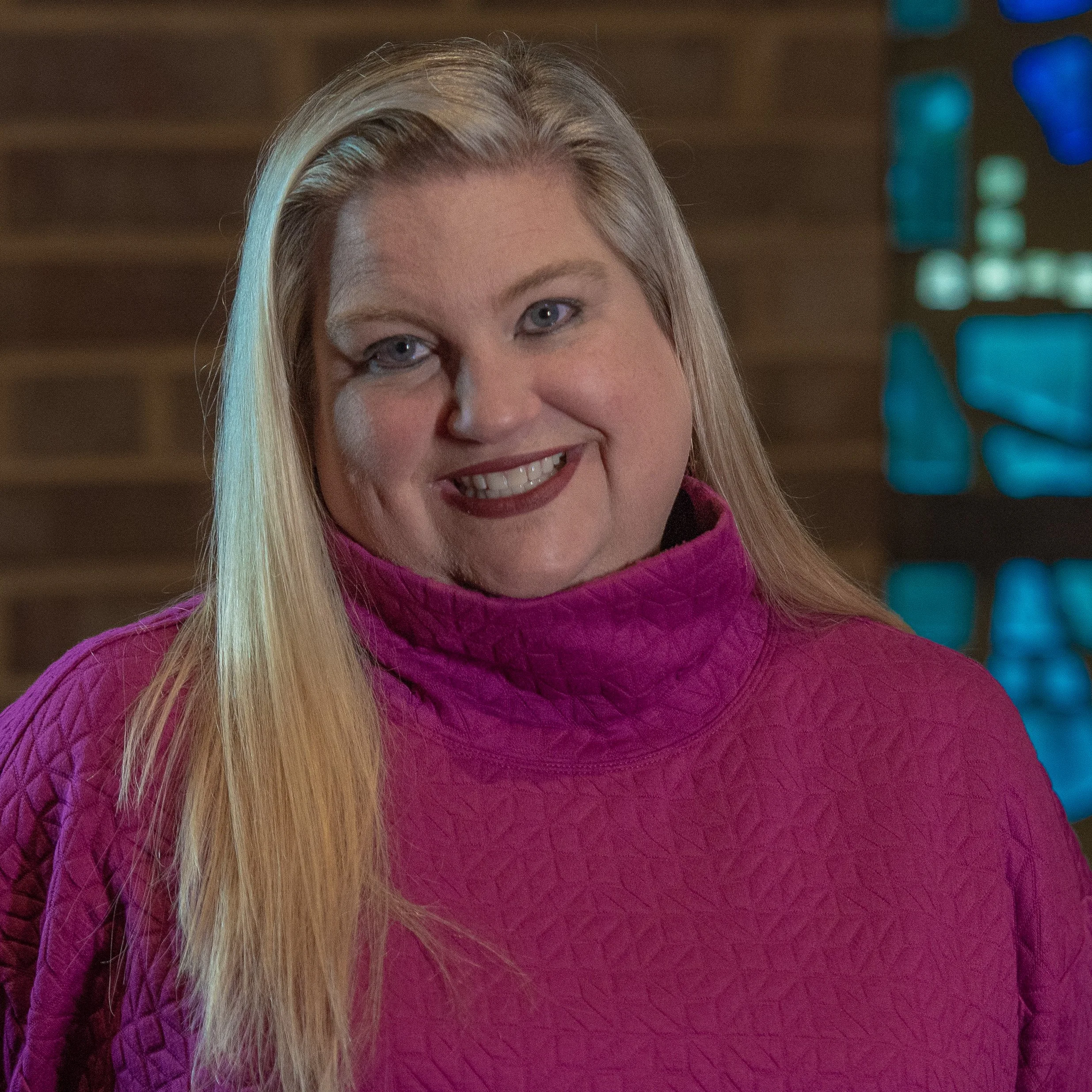 Woman with long blonde hair in pink quilted sweater, smiling, against a dark brick wall and blue stained glass background.