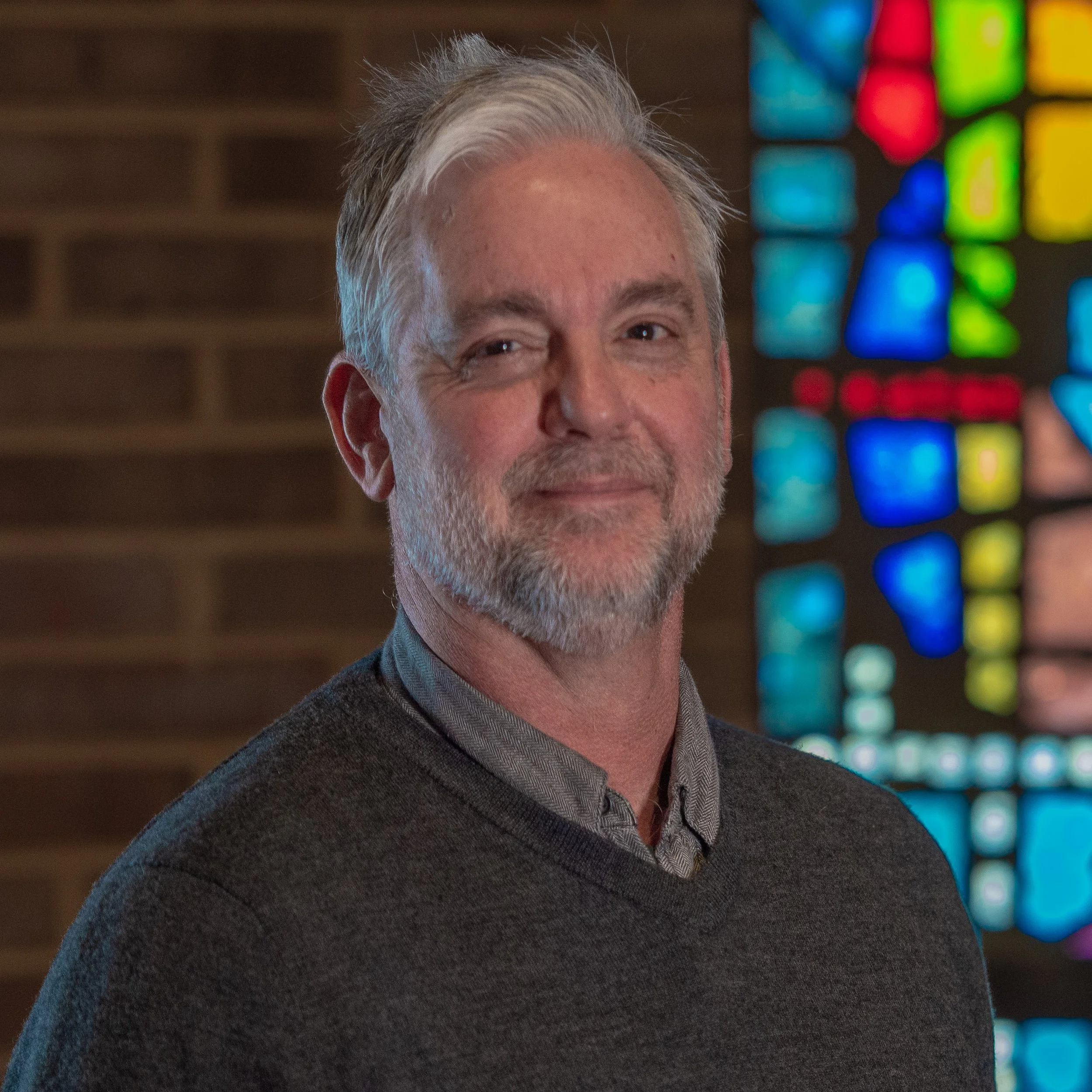 Man with gray hair and beard wearing a dark sweater standing indoors with a stained glass window in the background.