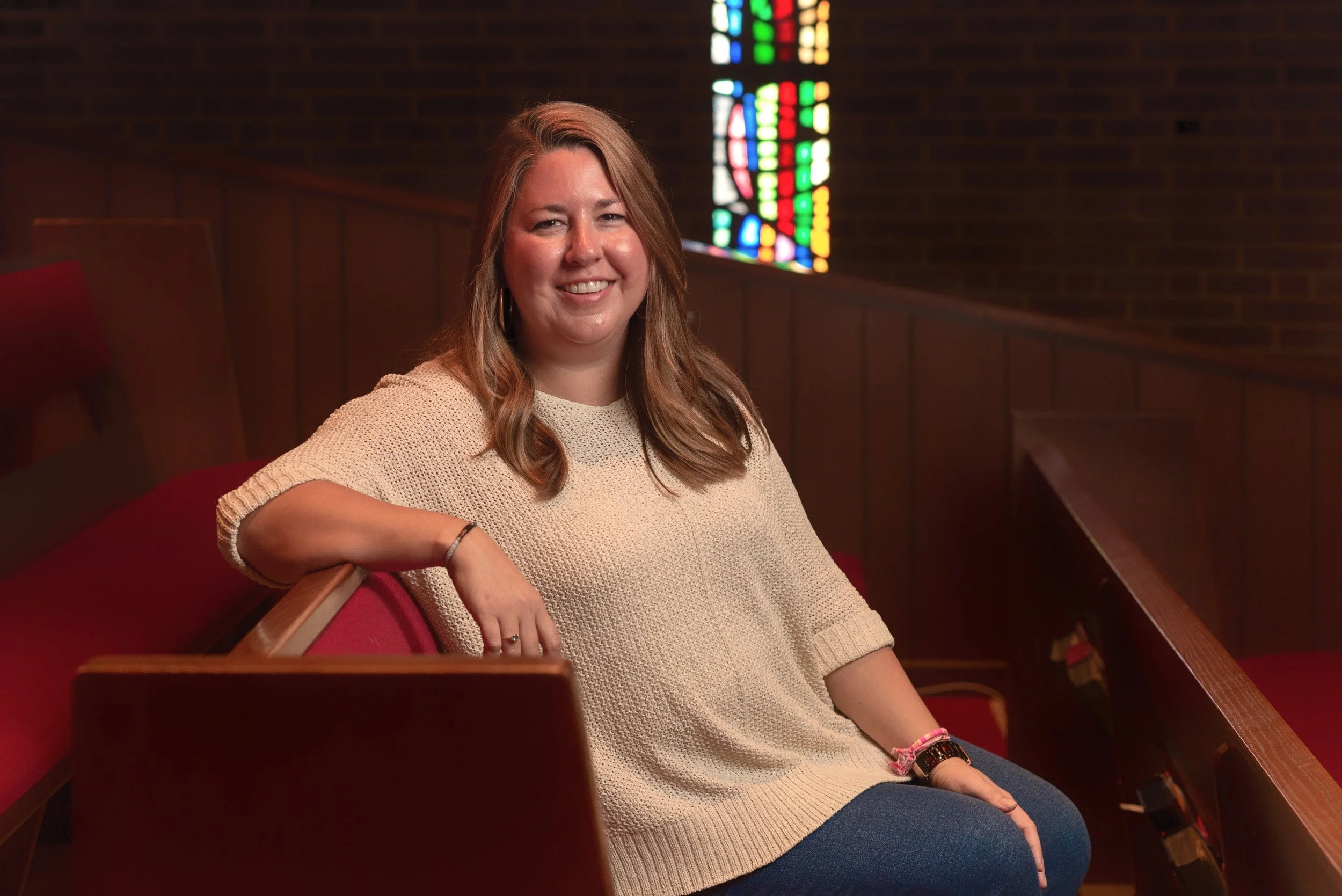 Woman smiling in church bench with stained glass window background.