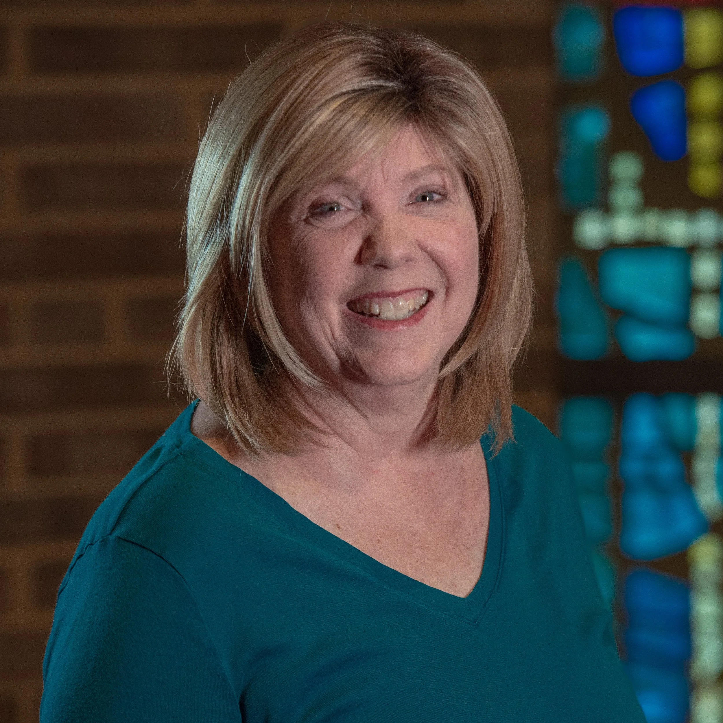 Smiling woman with blond hair in a teal shirt, standing in front of a stained glass window.