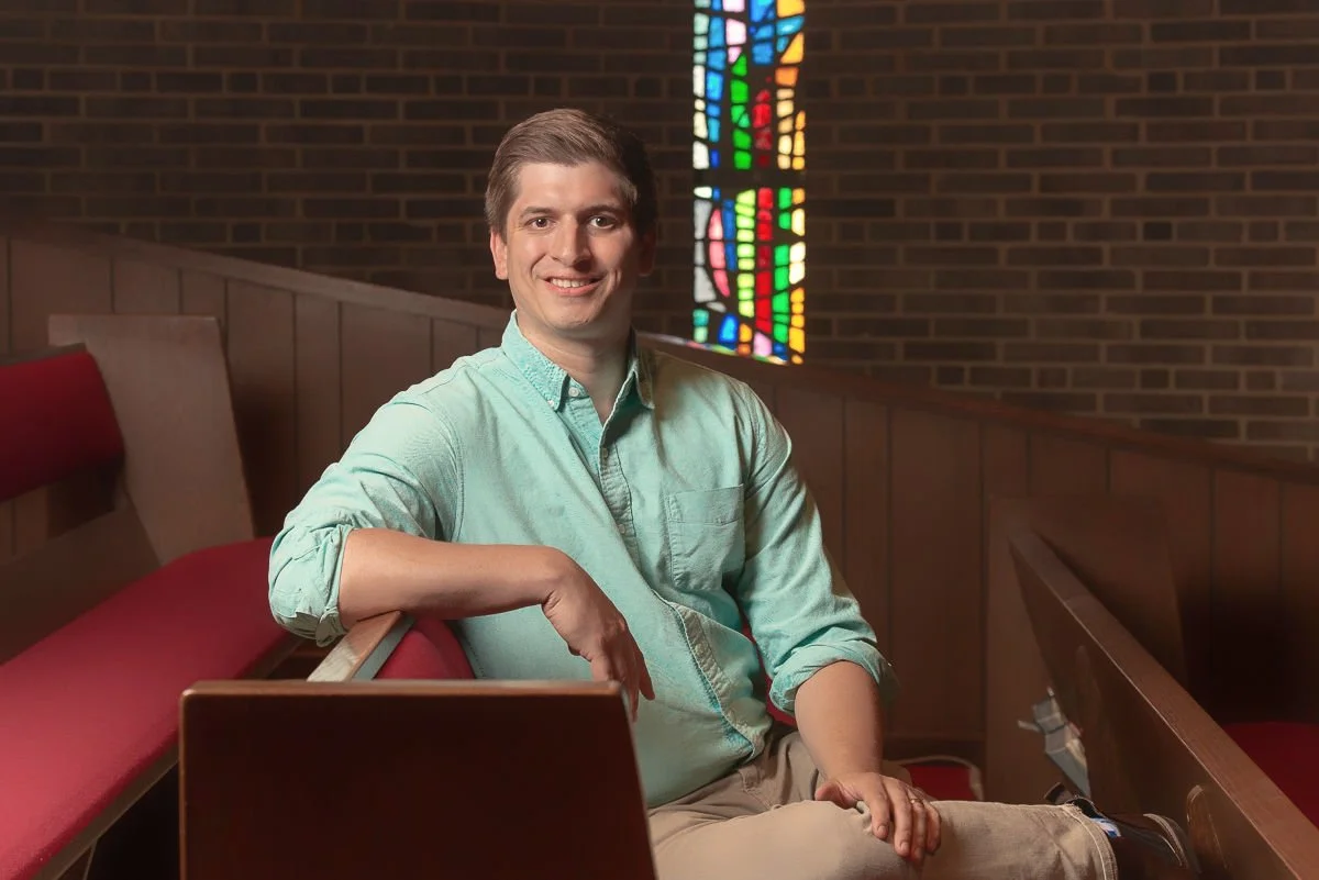 Man sitting in a church pew with stained glass window in the background.