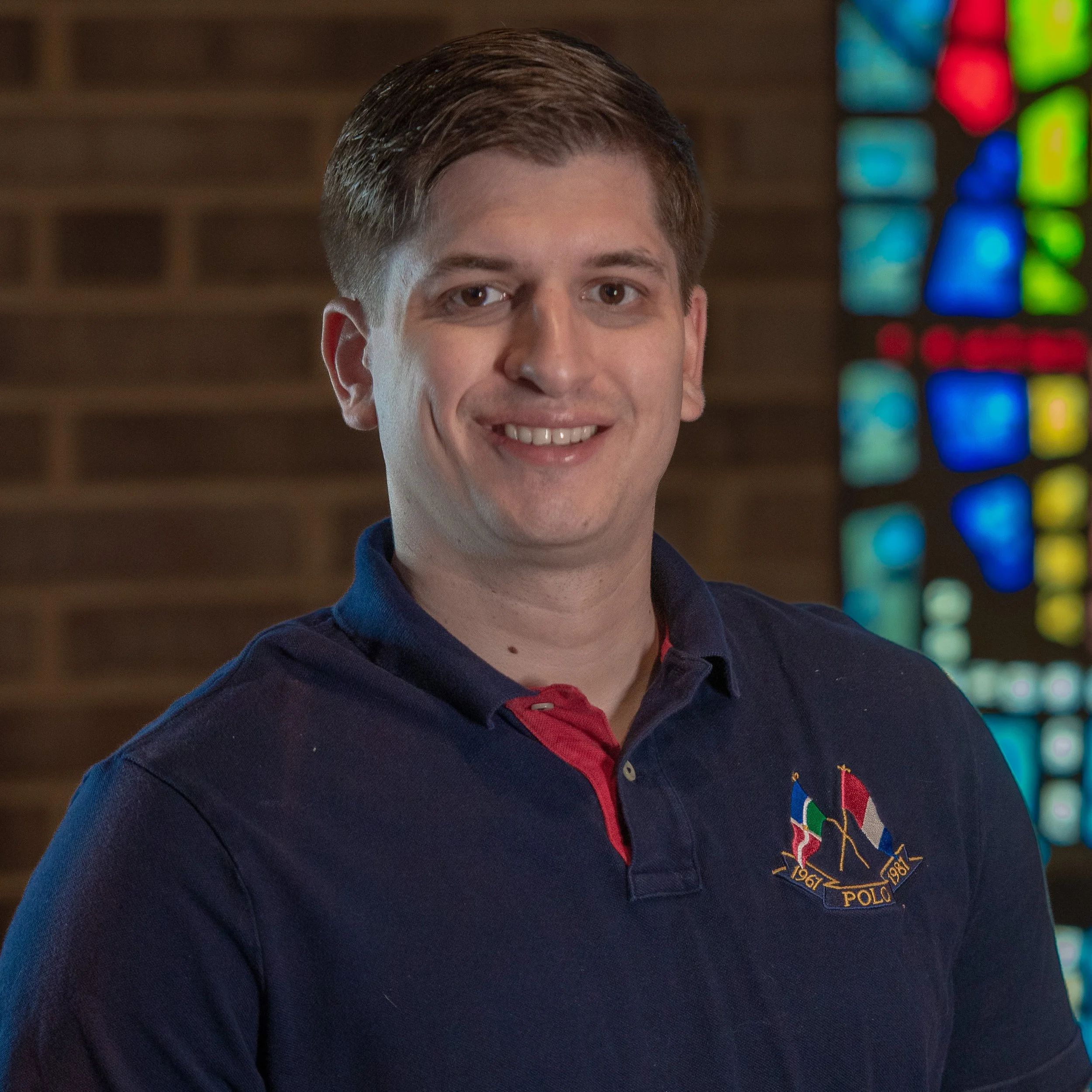 Person in a blue polo shirt standing indoors in front of a brick wall and stained glass window.