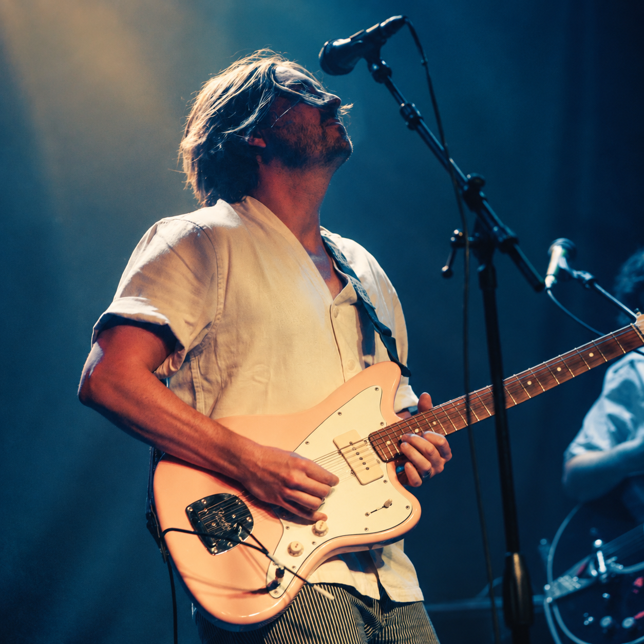 A man, Tyson Motsenbocker, playing an electric guitar on stage, with a microphone stand nearby, under warm stage lighting.