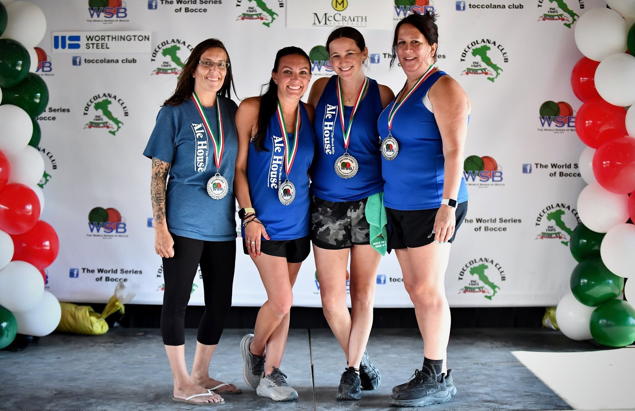 Four women on a podium at a bocce competition, wearing medals and athletic clothing, smiling for a photo, with a backdrop displaying Bocce event logos and colored balloons.