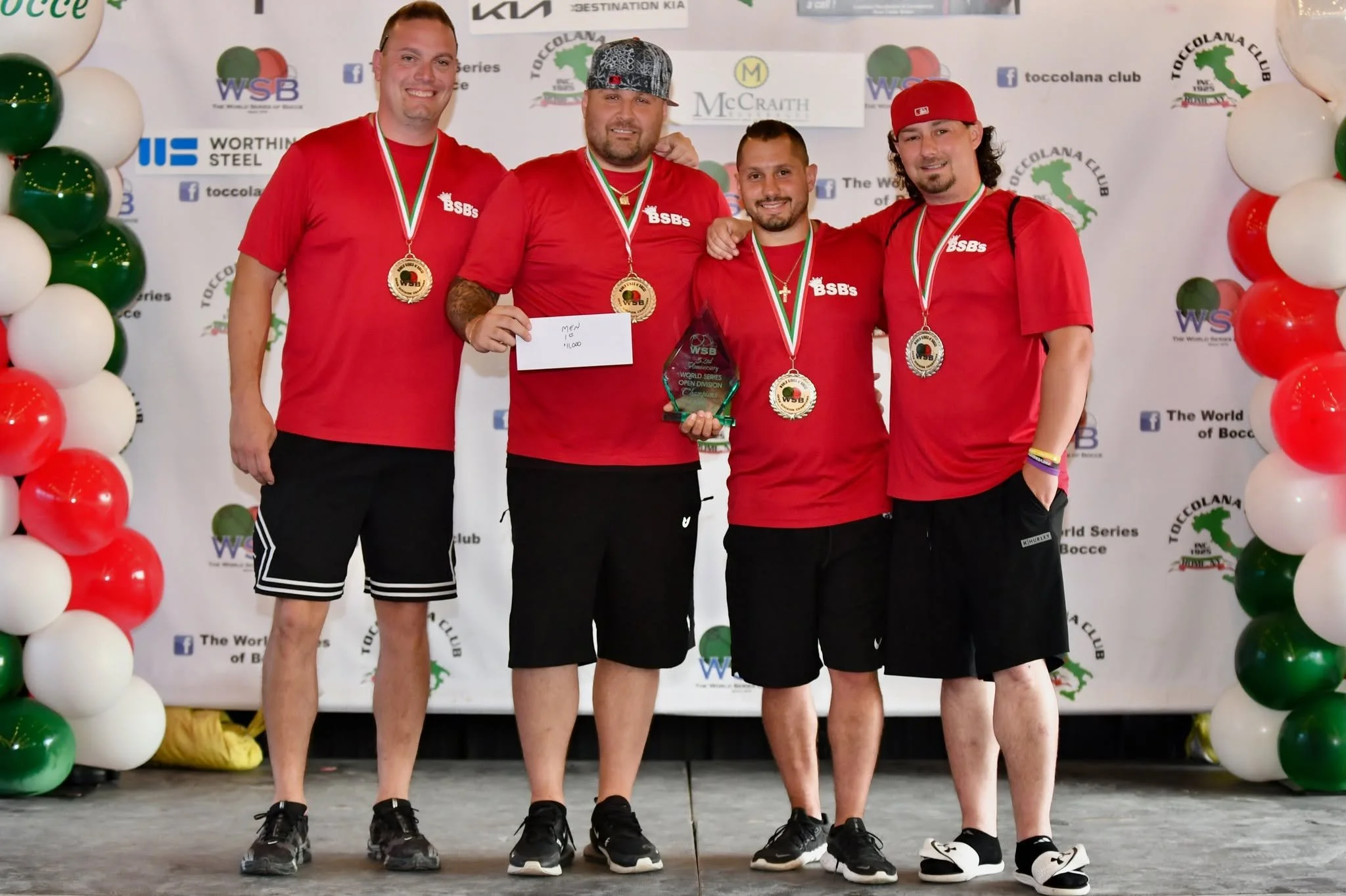 Four men wearing red bocce shirts and medals around their necks pose on a stage, holding a bocce tournament trophy and a check, in front of a backdrop with balloons and various logos.
