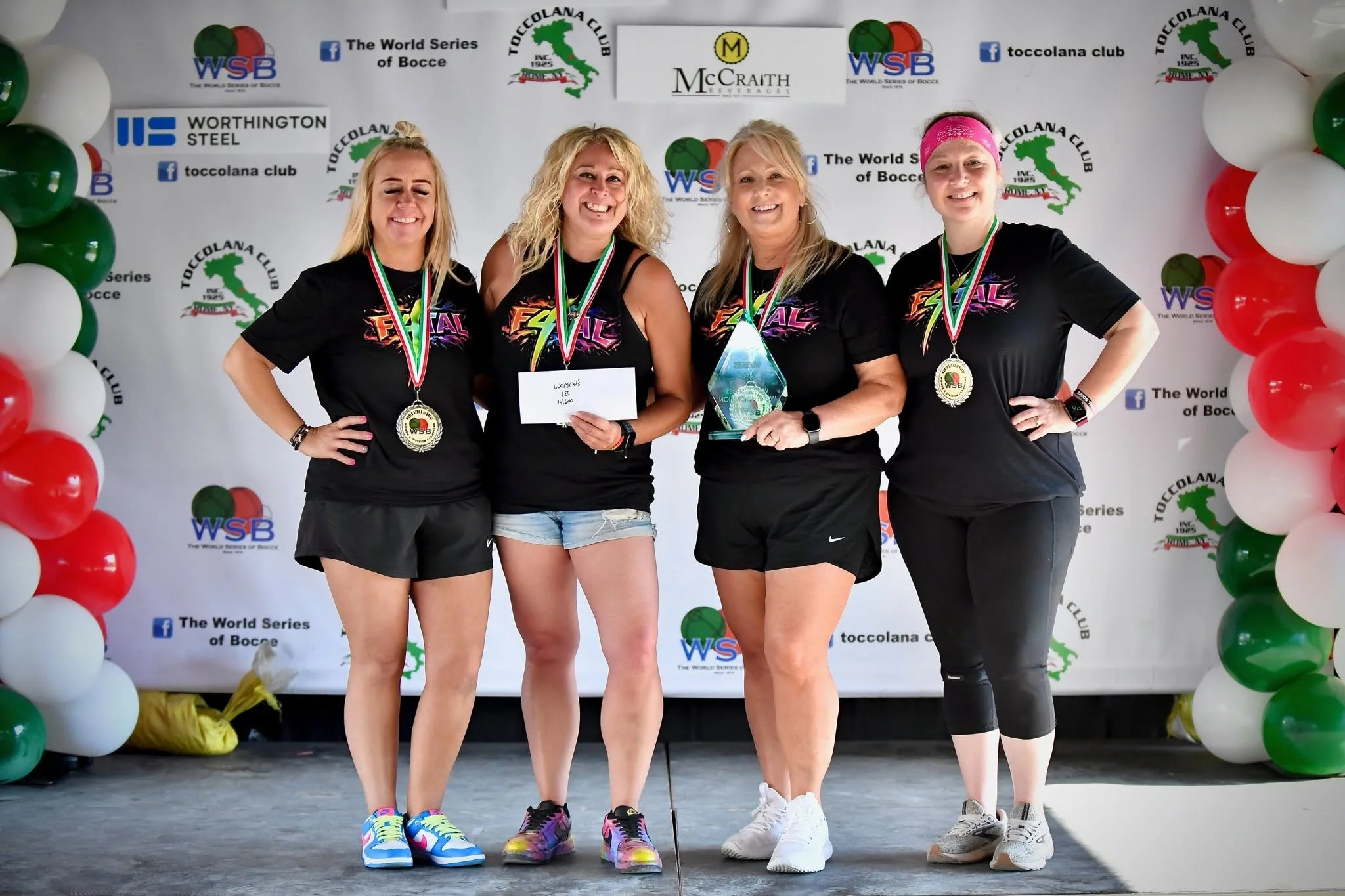 Four women standing on a stage with victory medals around their necks, smiling after a bocce tournament, with a backdrop featuring balloons and sponsorship logos.